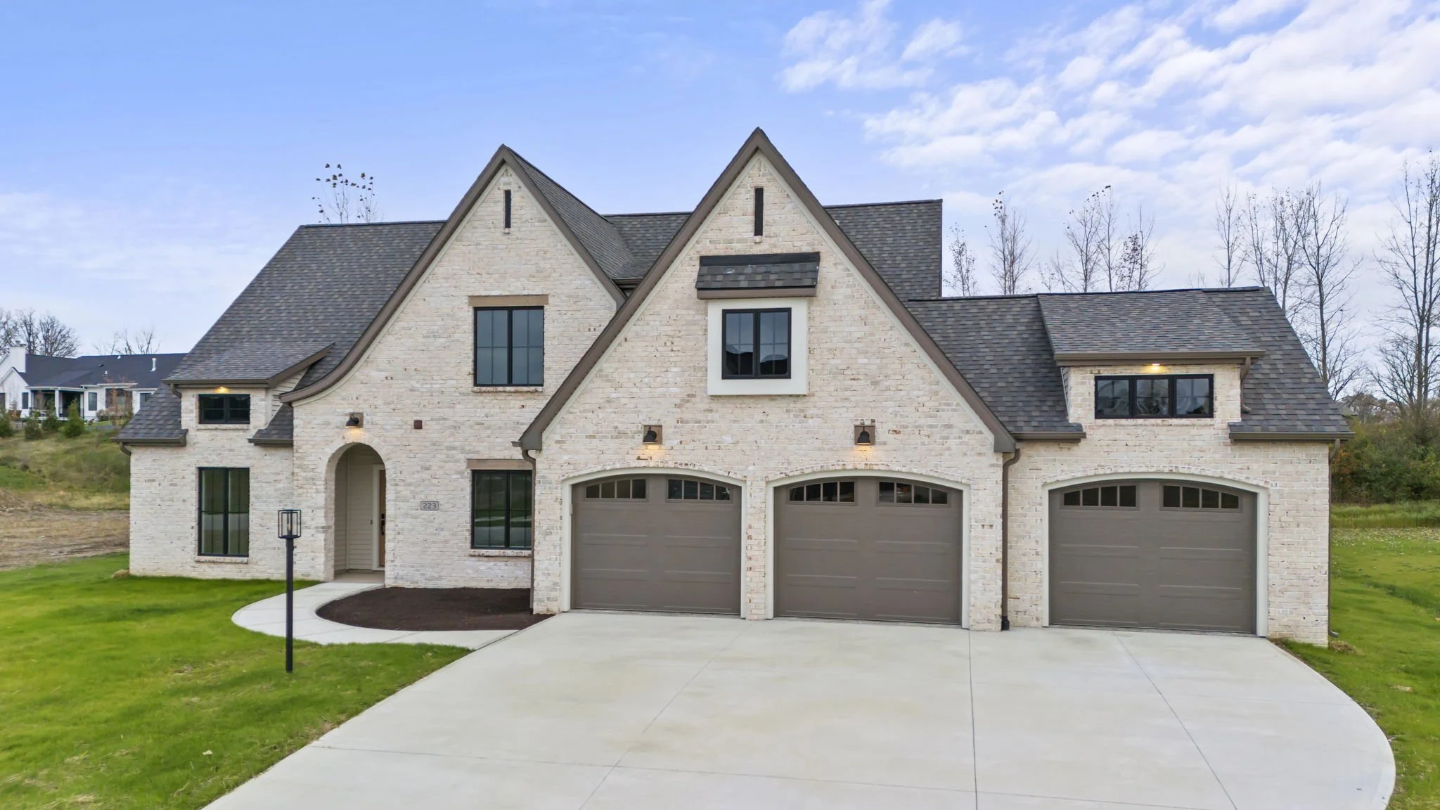 Front view of a modern white brick house with three garage doors, large windows, and a landscaped front yard under a partly cloudy sky.