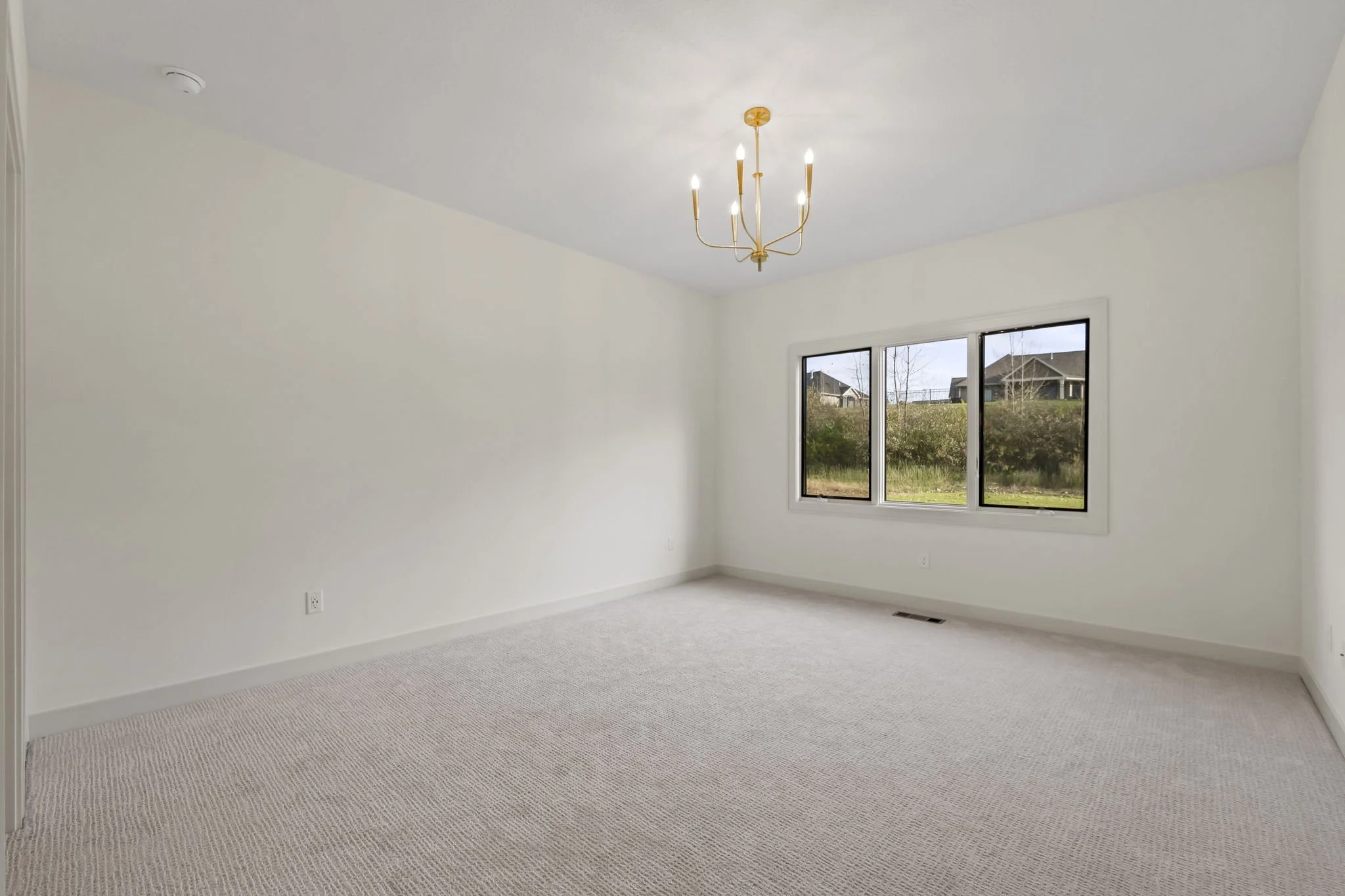 Empty room with white walls, beige carpet, a window showing houses outside, and a modern gold chandelier hanging from the ceiling.