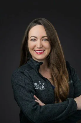 A woman with long brown hair smiling, wearing a dark jacket over a black shirt with a logo, against a dark background.
