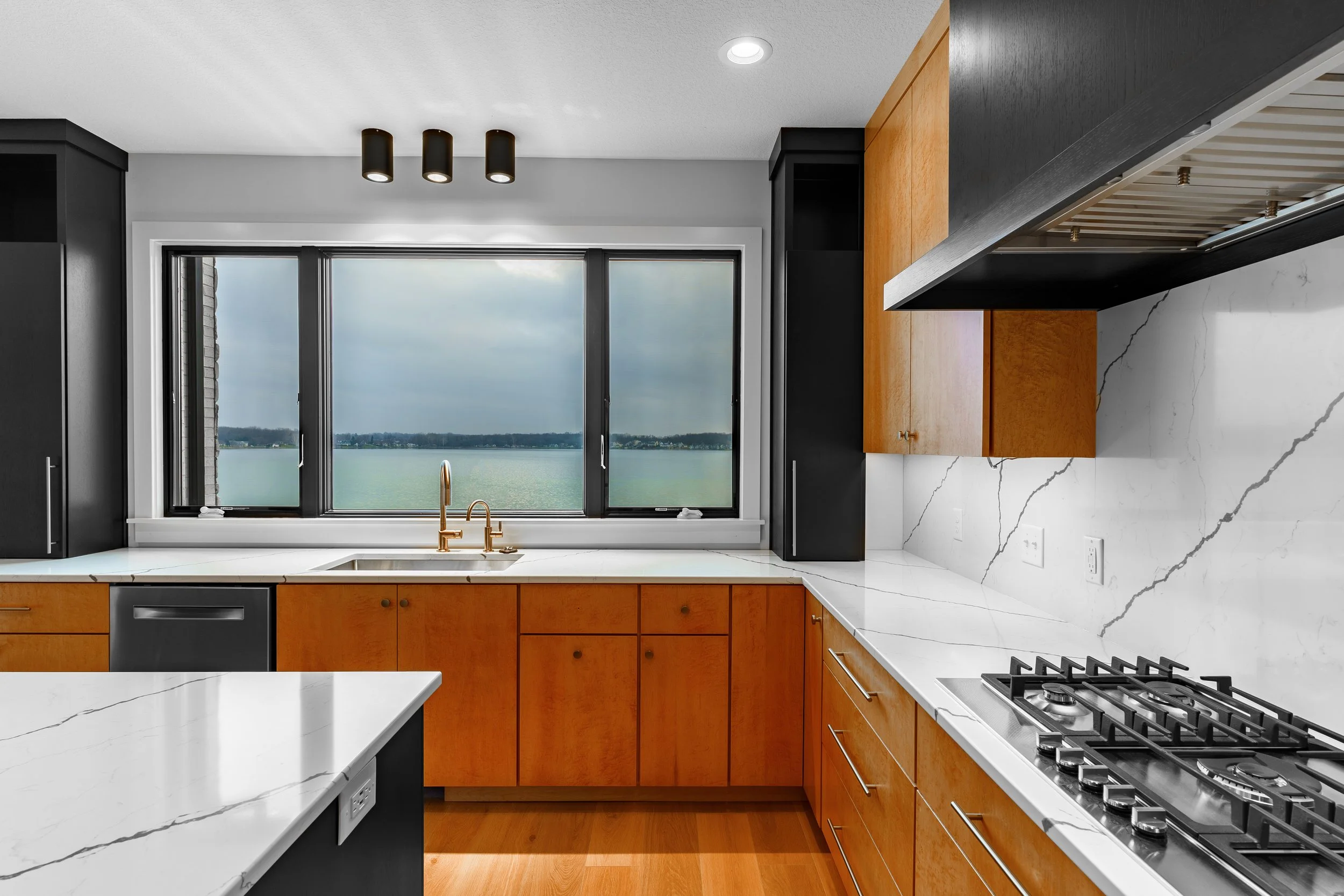 Modern kitchen with white marble countertops, wooden cabinets, black appliances, and a large window overlooking a body of water.