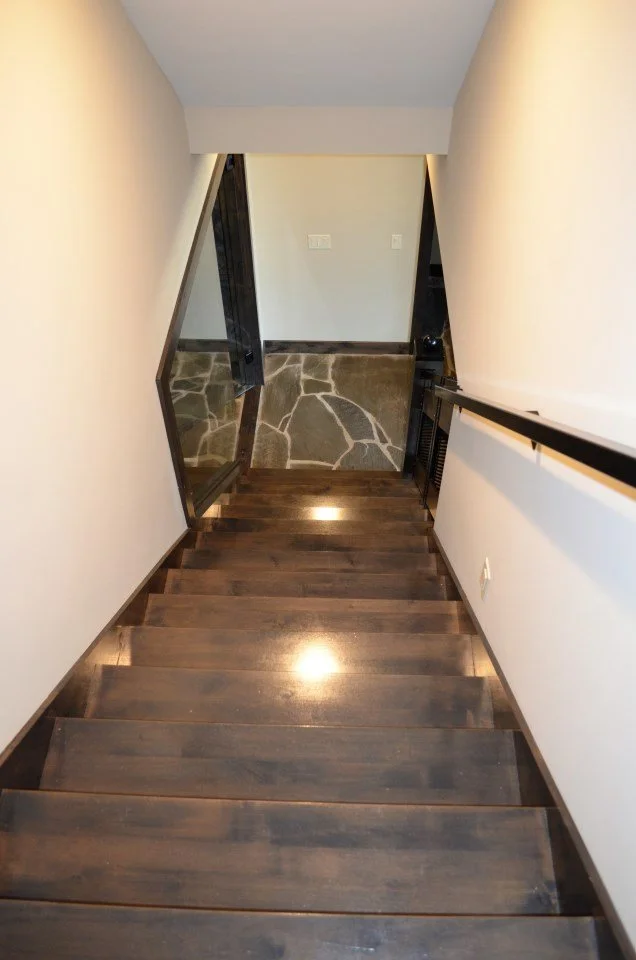 Wooden staircase leading down to a lower level, with a black handrail on the right side, beige walls, a stone-tile floor section at the bottom, and a doorway to the left.