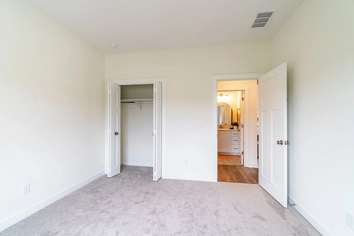 Empty bedroom with beige carpet, closet with bi-fold doors, and bathroom with vanity and mirror visible through open door.