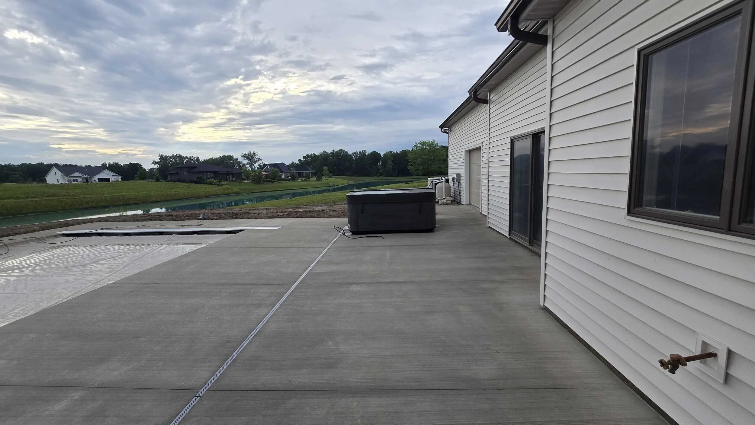 Backyard patio with concrete flooring, hot tub, and nearby hose bib, white house with sliding glass door, windows, and neighboring houses with lawns in the background under cloudy sky.