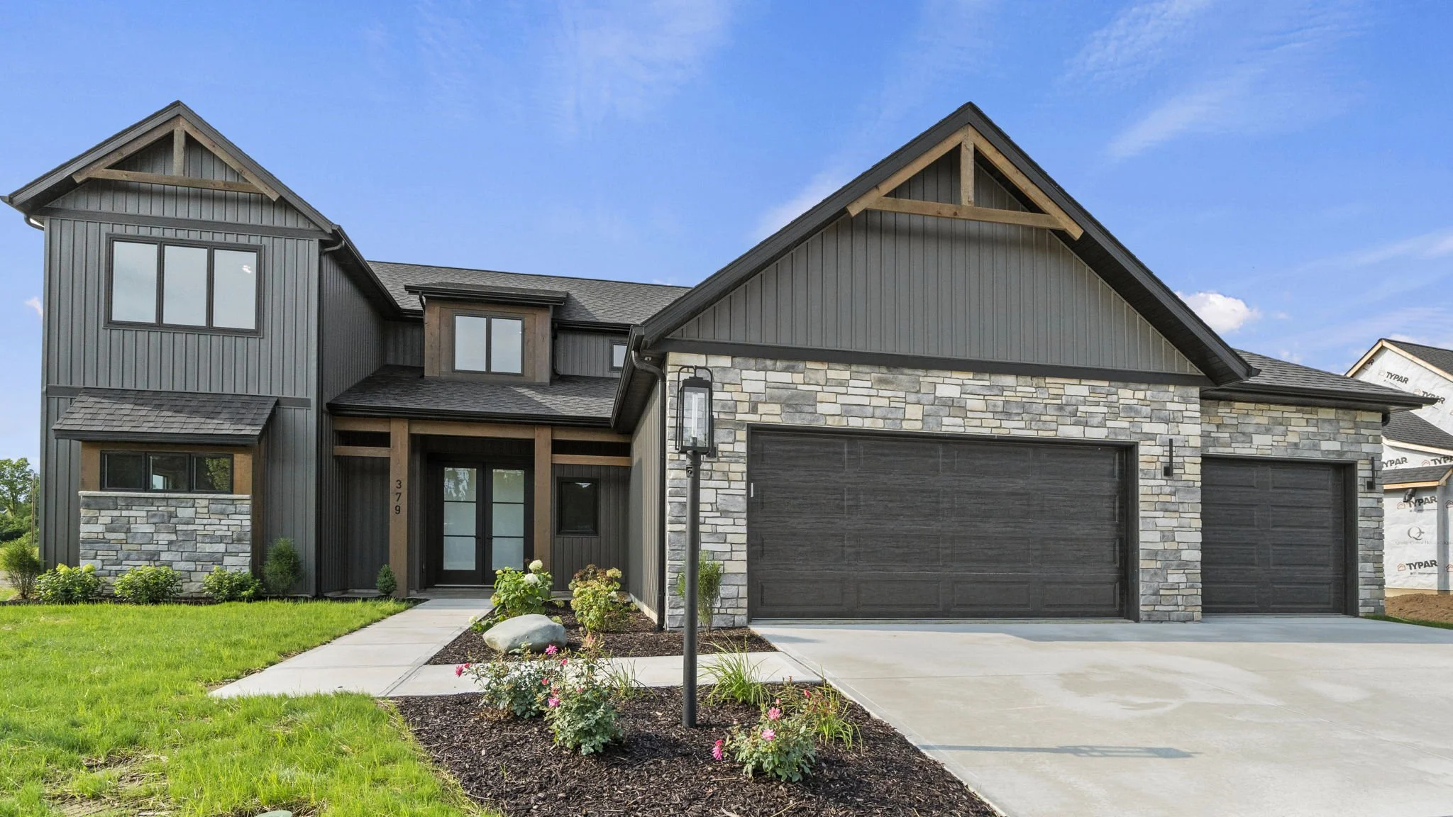 Modern two-story house with gray and black exterior, stone accents, and a front lawn with a garden. The house features a large double garage and front door with sidelight windows, and a lamp post near the walkway.