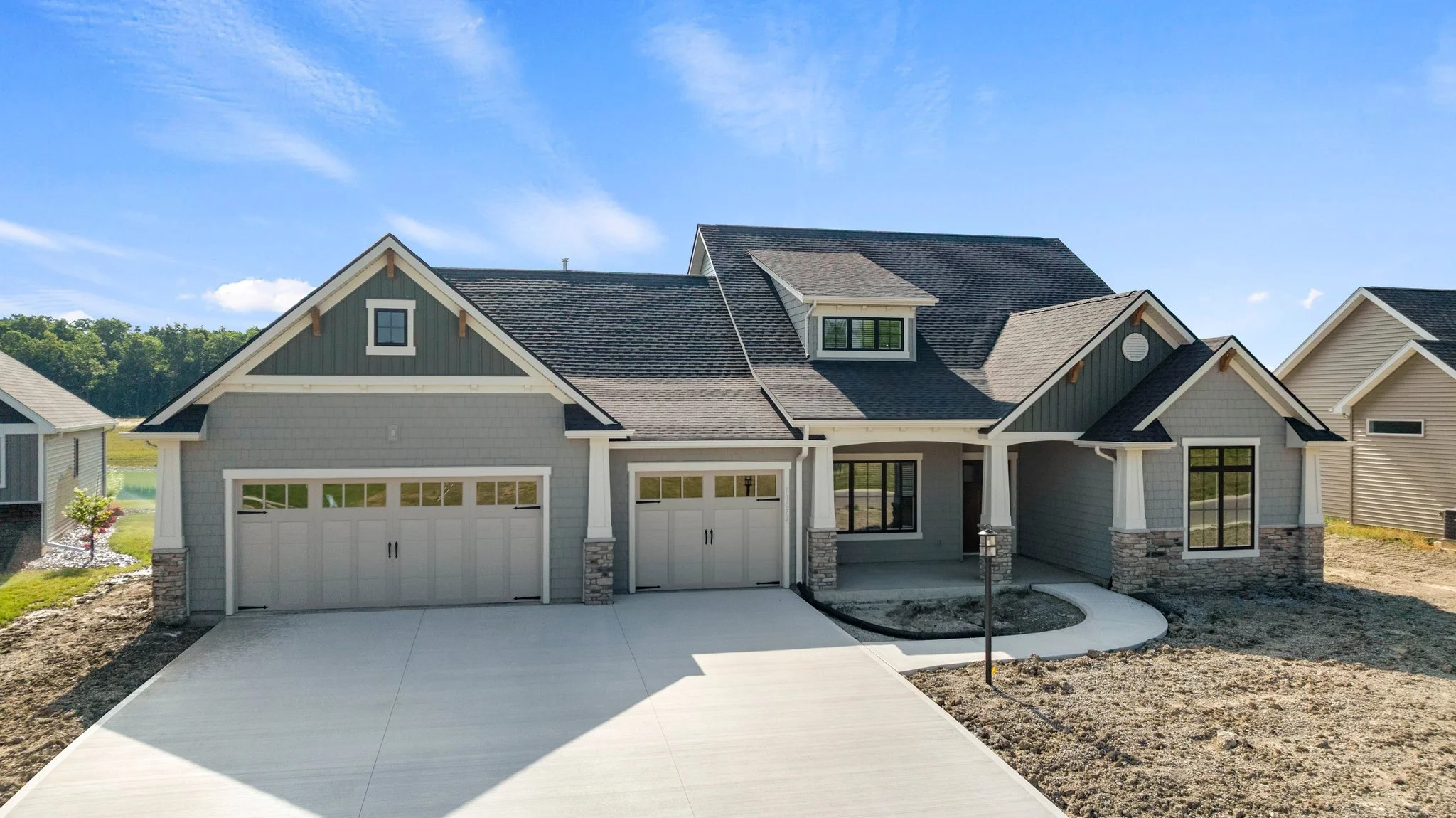 Front view of a new two-story house with gray siding, stone accents, and a concrete driveway, under a blue sky.