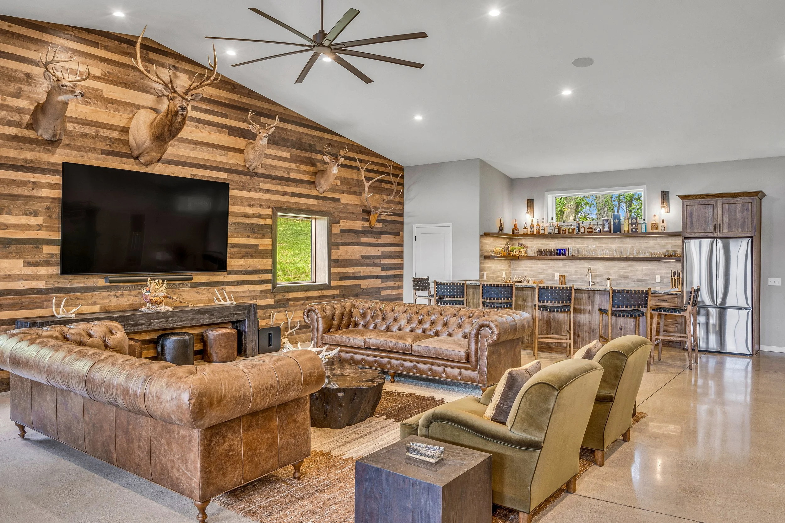 Living room with wall decorated with mounted deer heads, a large flat-screen TV, and a bar area with barstools in the background.