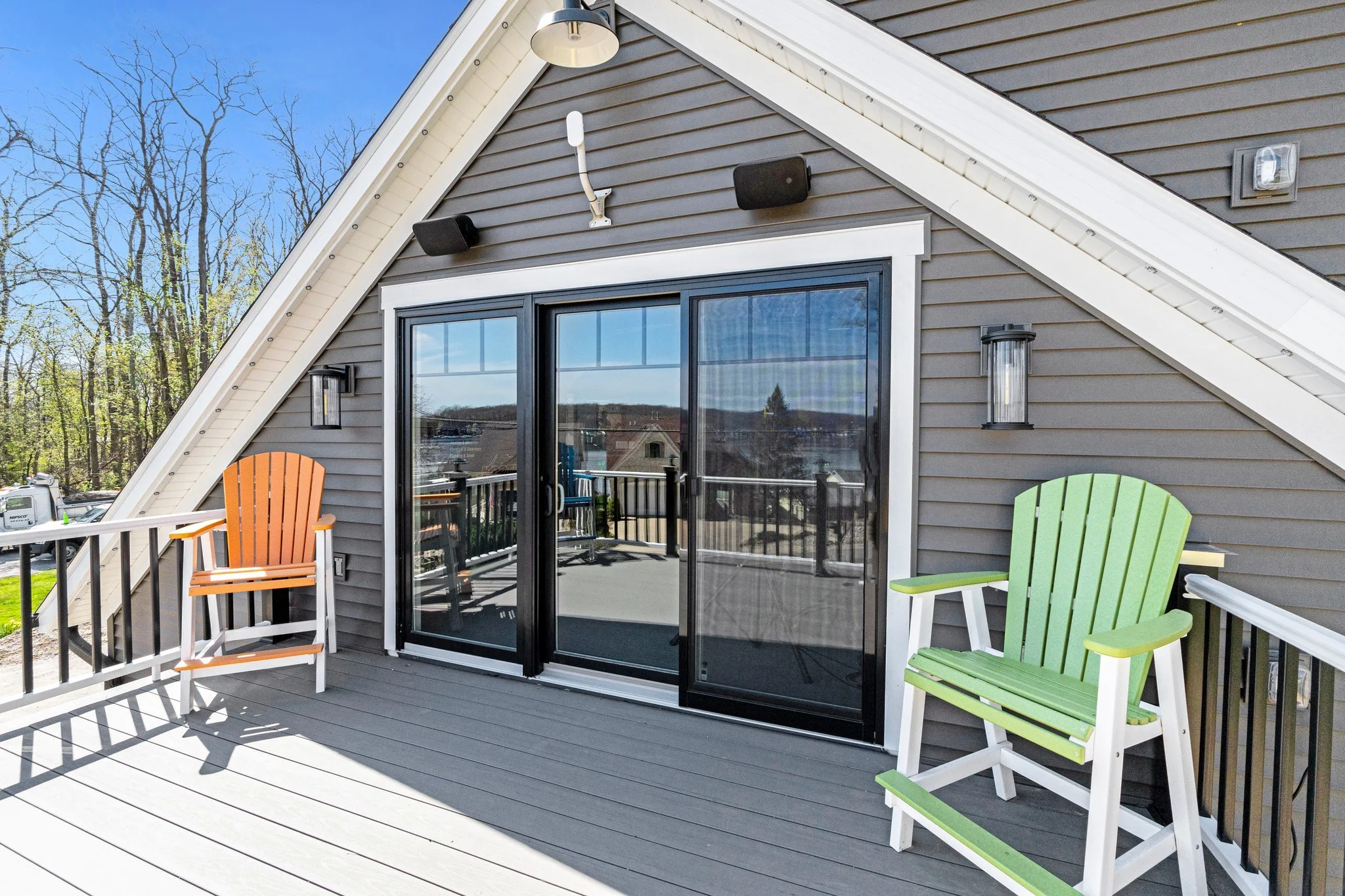 Balcony with sliding glass door, two Adirondack chairs in orange and green, outdoor wall lights, and an exterior wall with gray siding and outdoor lights.