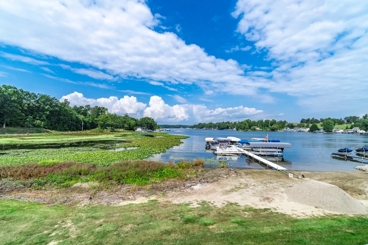 A lakeside scene showing a blue sky with white clouds, green trees along the shoreline, and a dock with boats on calm water.