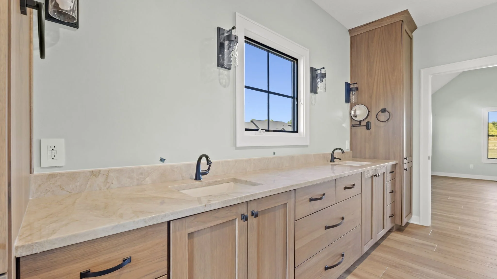 A modern kitchen with a beige marble countertop, wood cabinets, and two sinks with black faucets. There is a window above the countertop and bathroom-style wall sconces on either side. The room opens to a bright room with a large window and wood floo