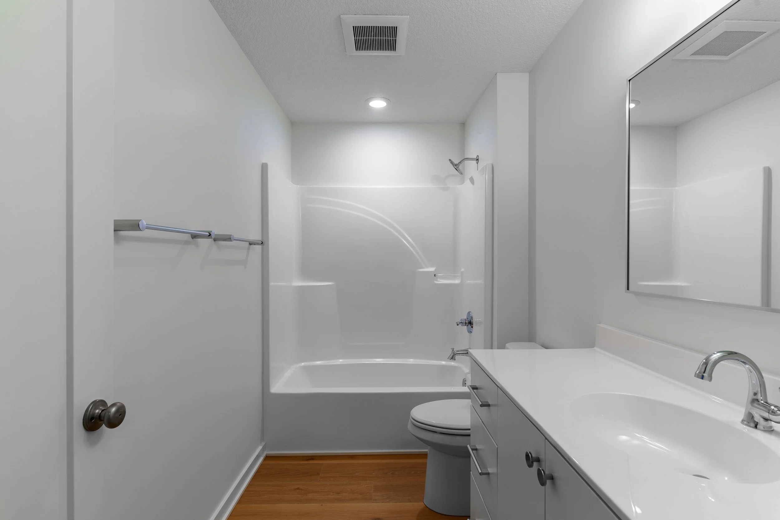A small, modern bathroom with a white bathtub, toilet, and a white vanity with a sink. There is a large mirror above the vanity, a towel bar on the wall, and a wood floor.