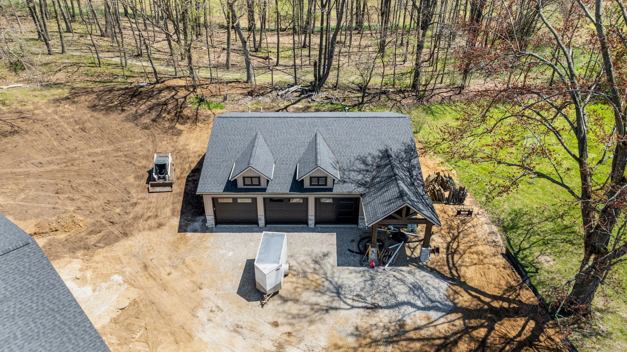 Aerial view of a house under construction with a dark grey roof, a gravel driveway, construction equipment, and surrounding trees and grassy area.