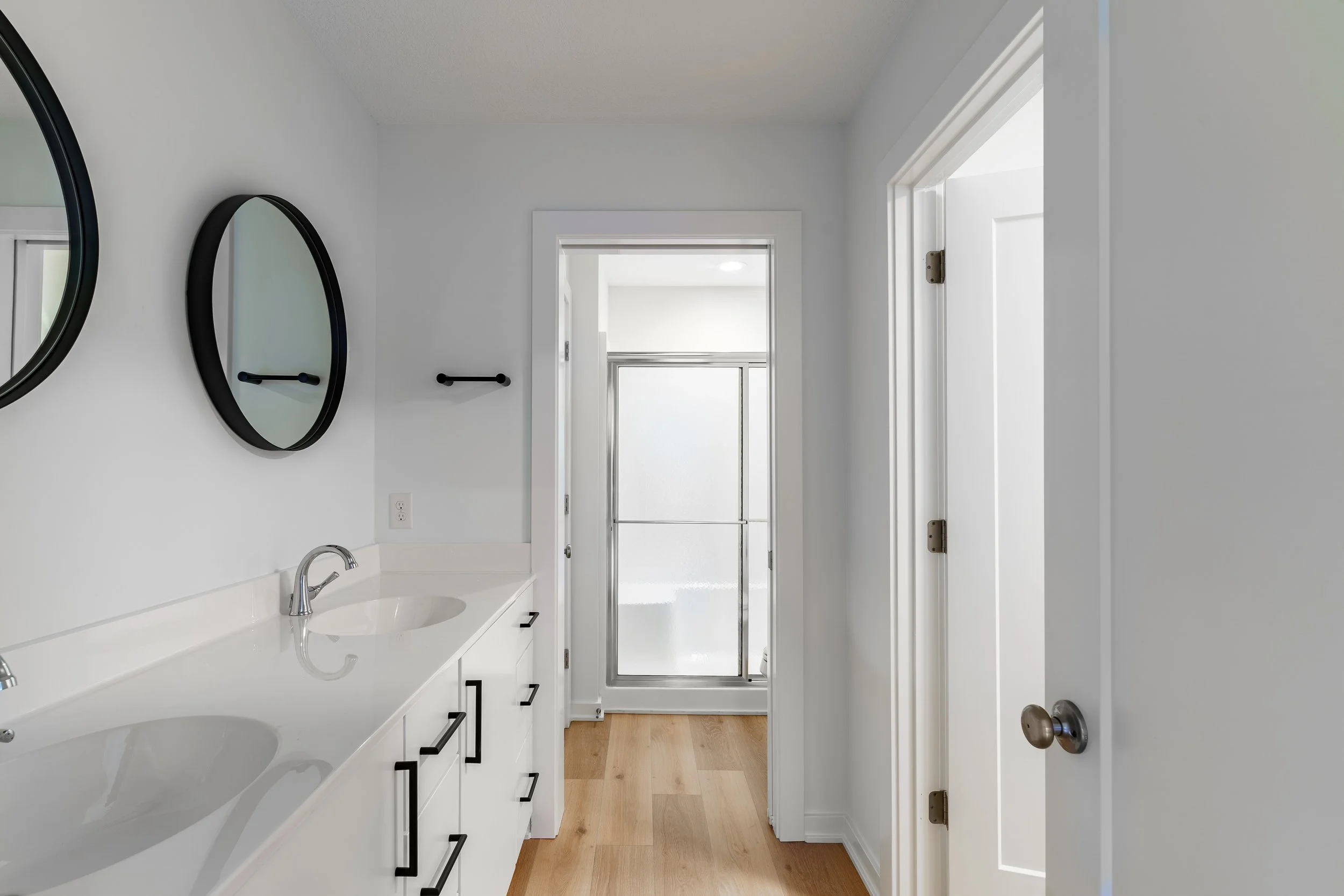 Modern bathroom with dual sinks, white vanity, black handles, round mirrors, and a frosted glass shower door at the end of the room.