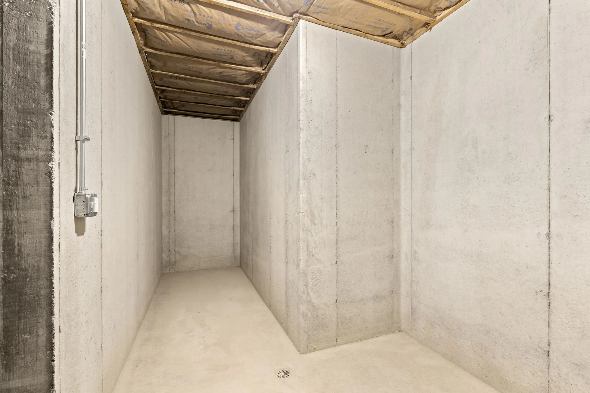 An empty unfinished room with bare concrete walls, floor, and ceiling with exposed insulation and electrical conduit.