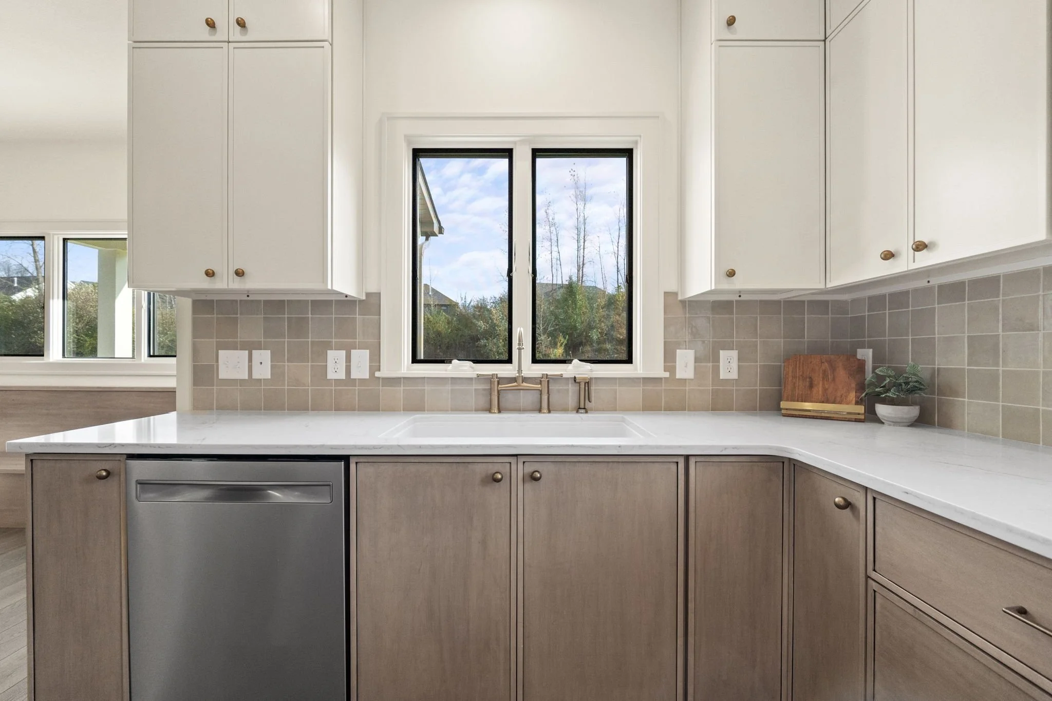 Kitchen sink area with white upper cabinets, light wood lower cabinets, beige tiled backsplash, a window over the sink showing an outdoor scene, and a small plant with cutting board on the counter.