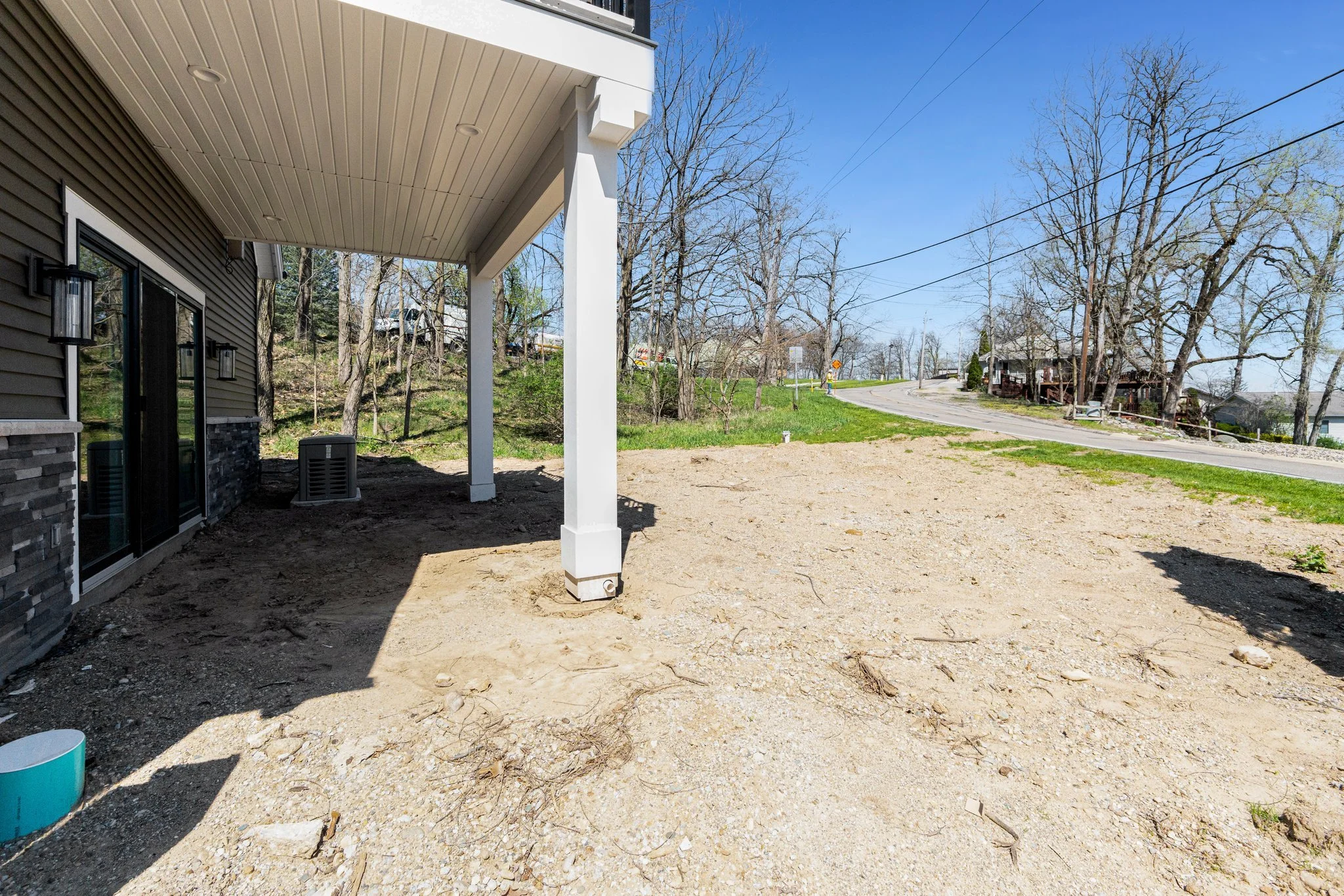 A backyard patio area under construction with exposed dirt and no grass, next to a house with sliding glass doors and exterior lights, overlooking a curved street with leafless trees and a few houses in the background.