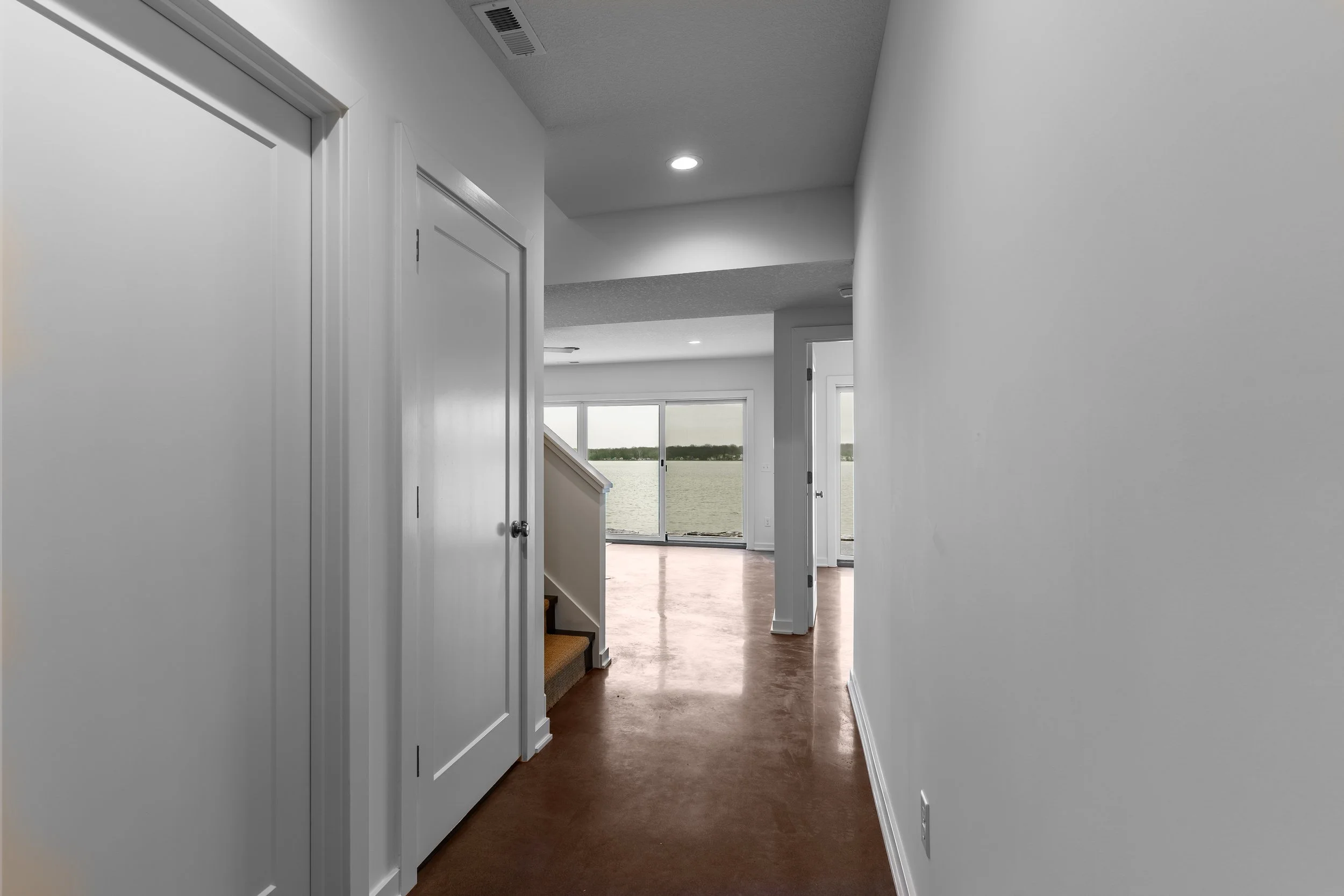 Empty hallway in a house with white walls, brown flooring, and a view of a large window overlooking a body of water.