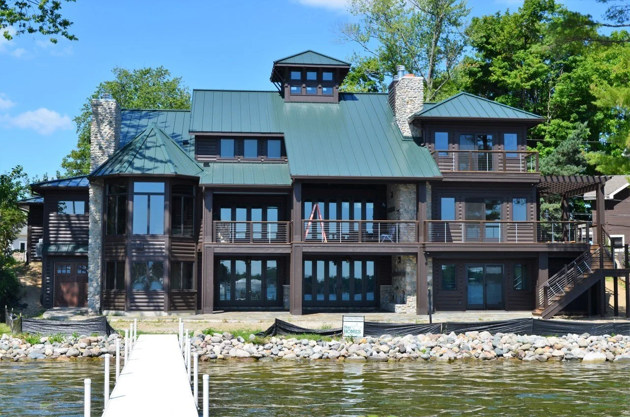 Large waterfront house with a dock, multiple balconies, and a green metal roof, surrounded by trees and rocks along a lake.