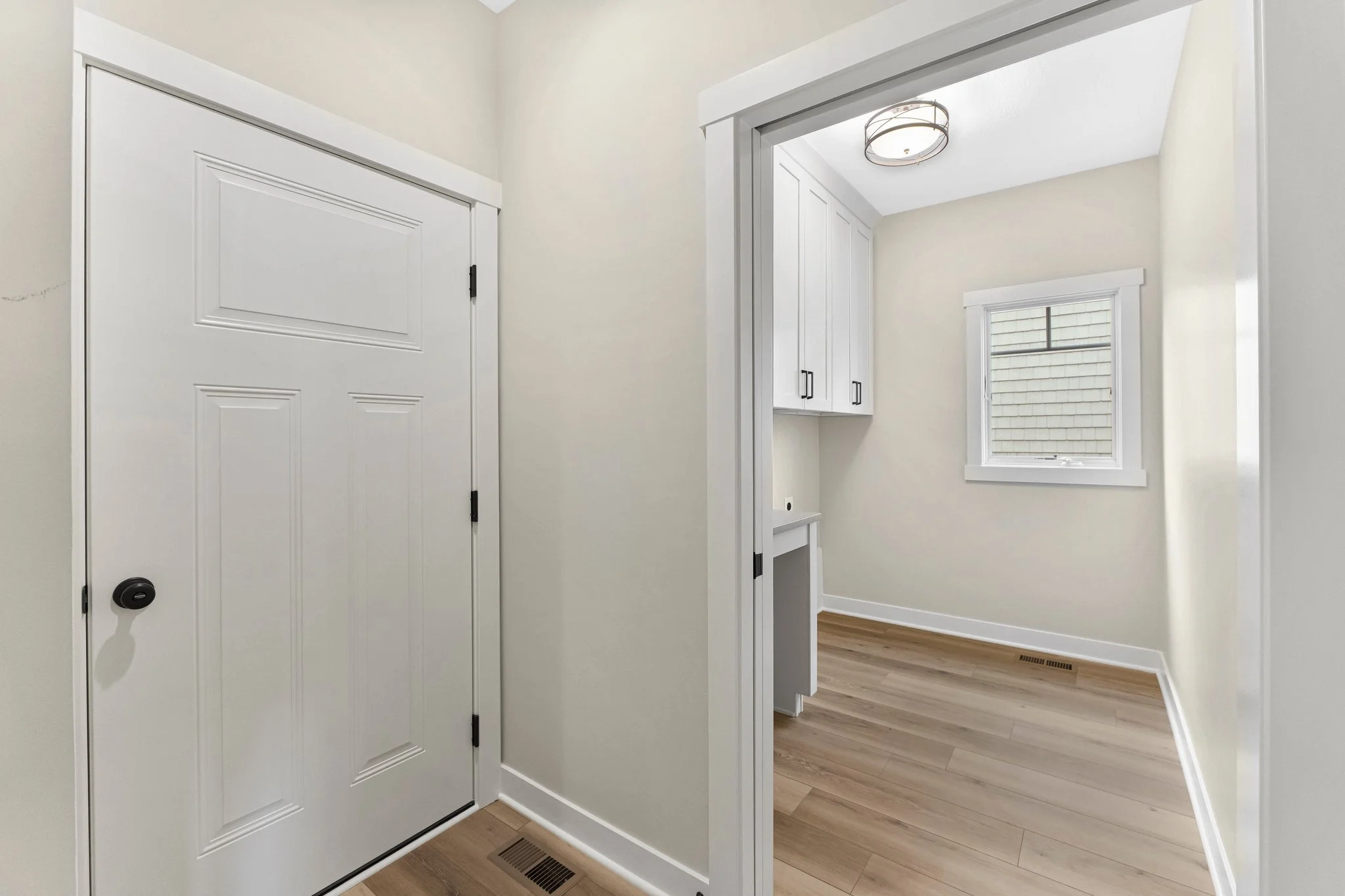 A small room with a window and white cabinets, seen through a doorway from an adjacent area with a white door and a vent on the wooden floor.