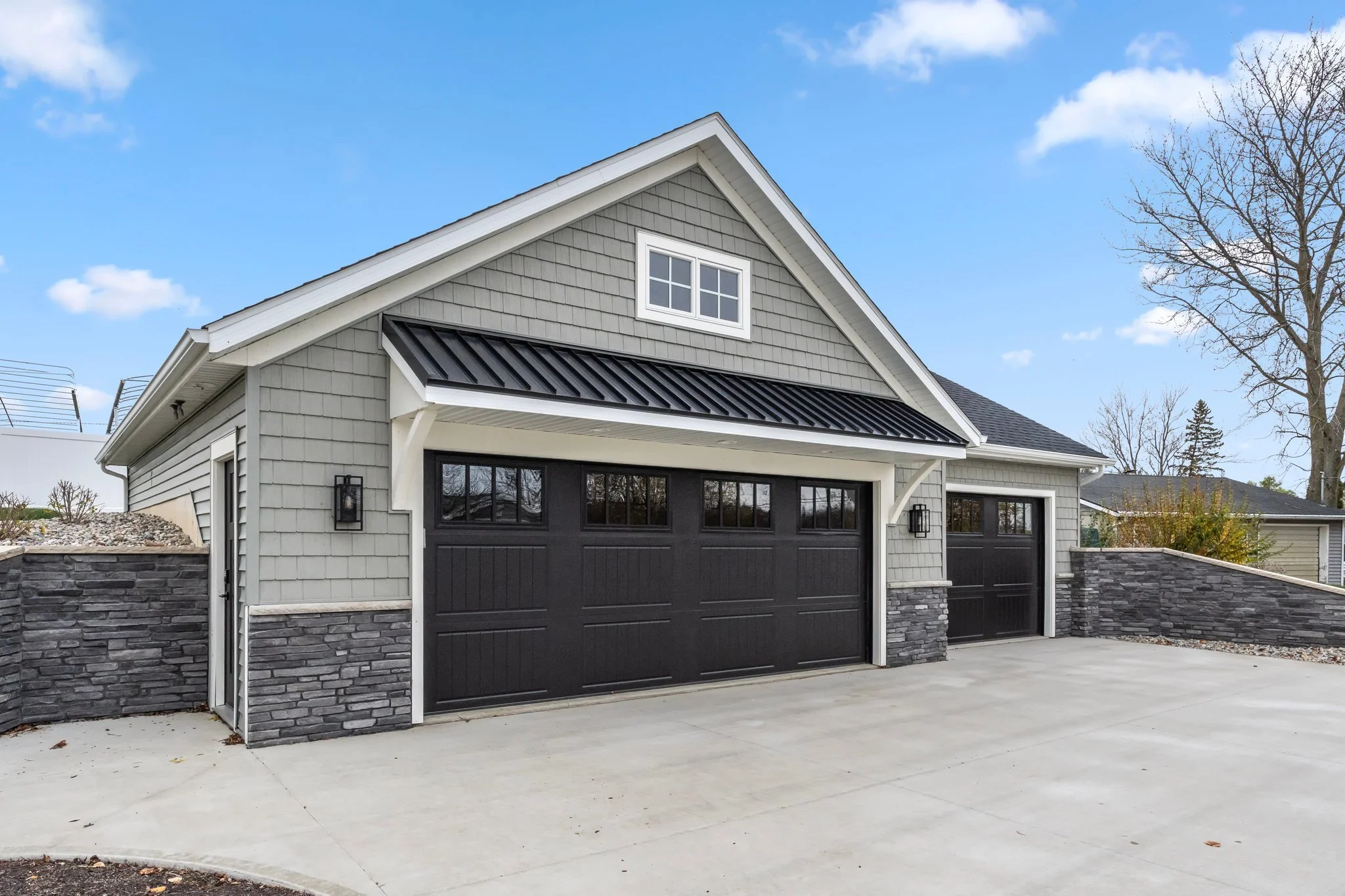 Modern house with black garage doors, gray exterior siding, stone accents, concrete driveway, and a blue sky with clouds.