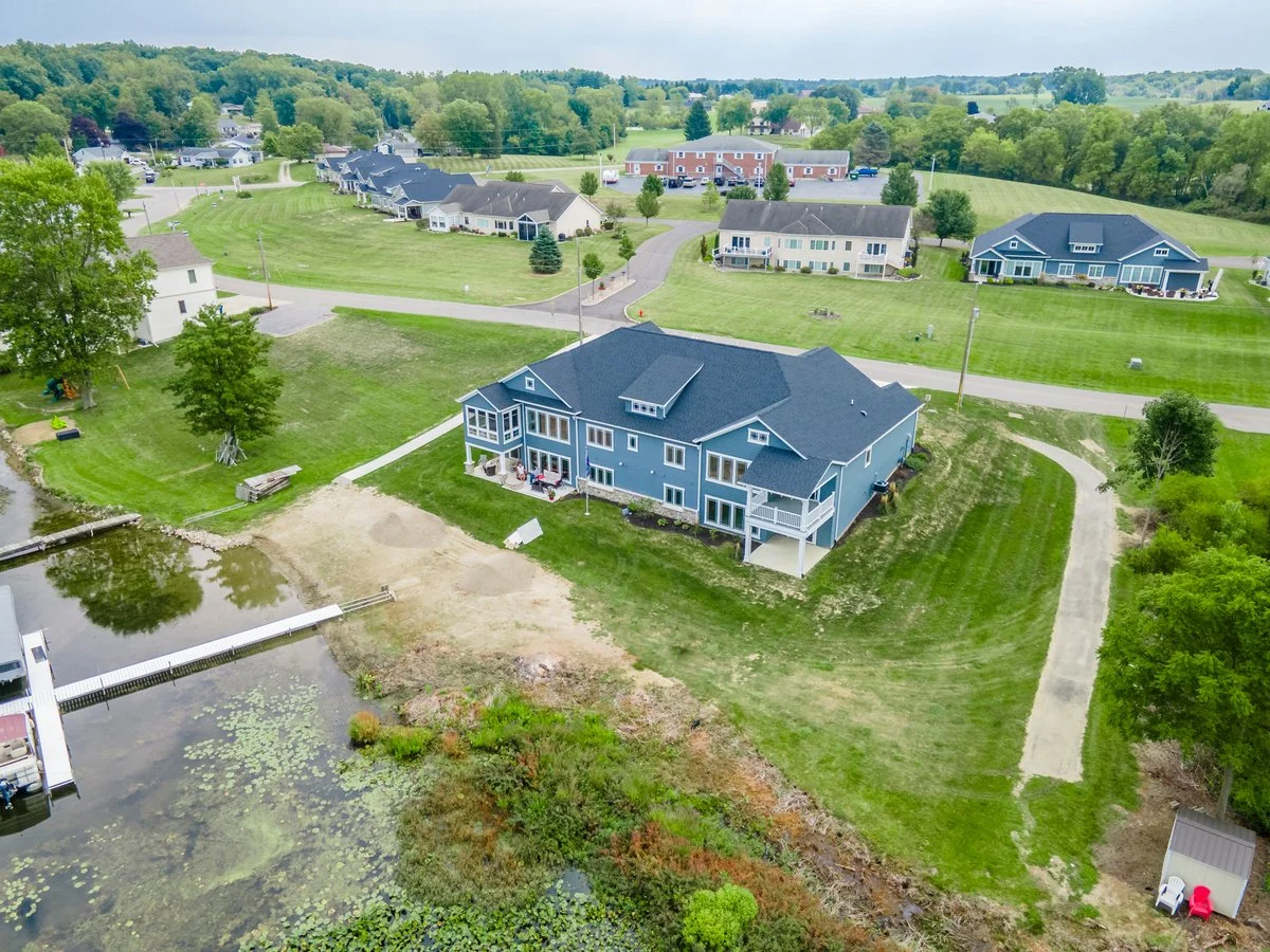 A large blue house by a pond with a dock, surrounded by green lawns and other houses in a suburban neighborhood. Trees line the area, and a gravel driveway is visible.