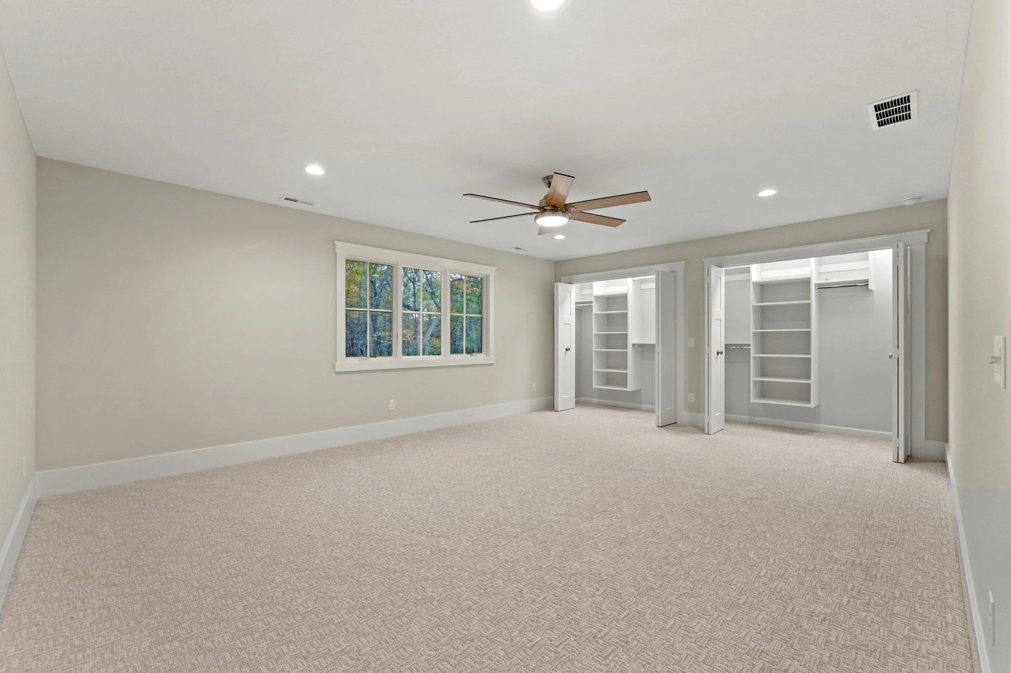 Empty bedroom with a beige carpet, white walls, a ceiling fan, three windows, and two built-in closets with open doors.
