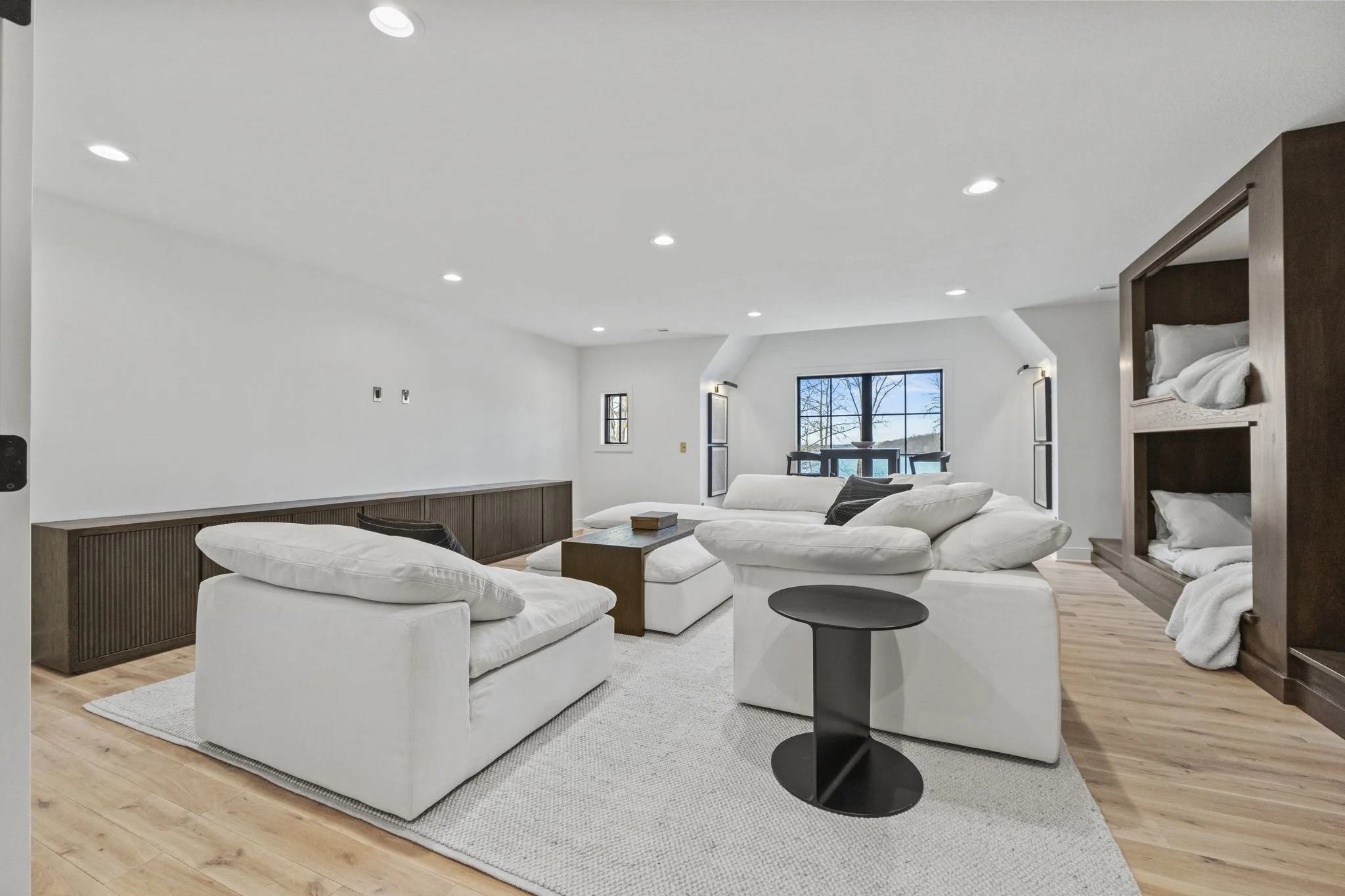 Living room with white sofas, black side table, and wooden flooring, featuring large windows with view outside.