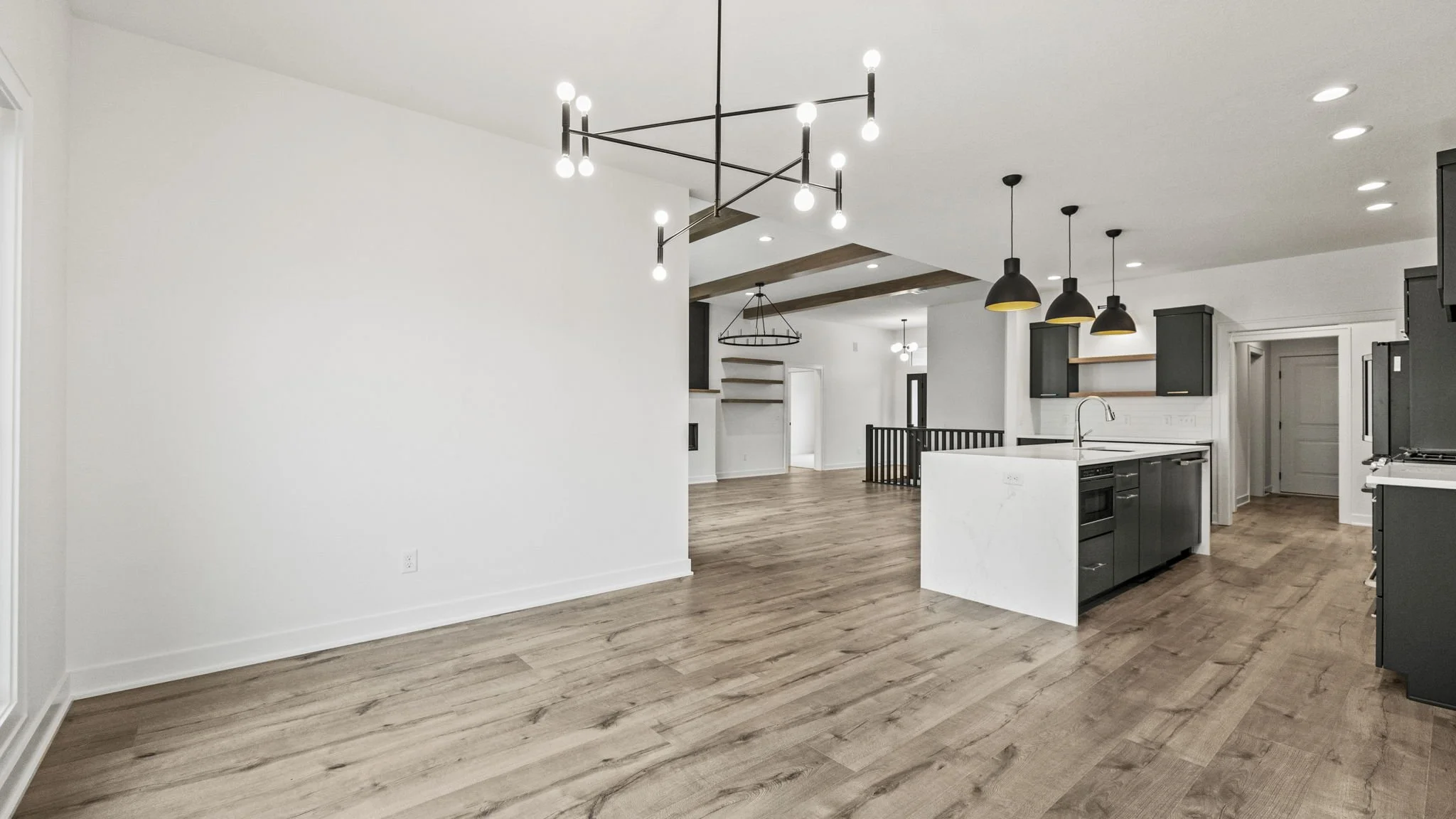 Open-concept kitchen and living area with white walls, wooden floors, black kitchen cabinets, a white marble island, black pendant lights, and modern lighting fixtures.