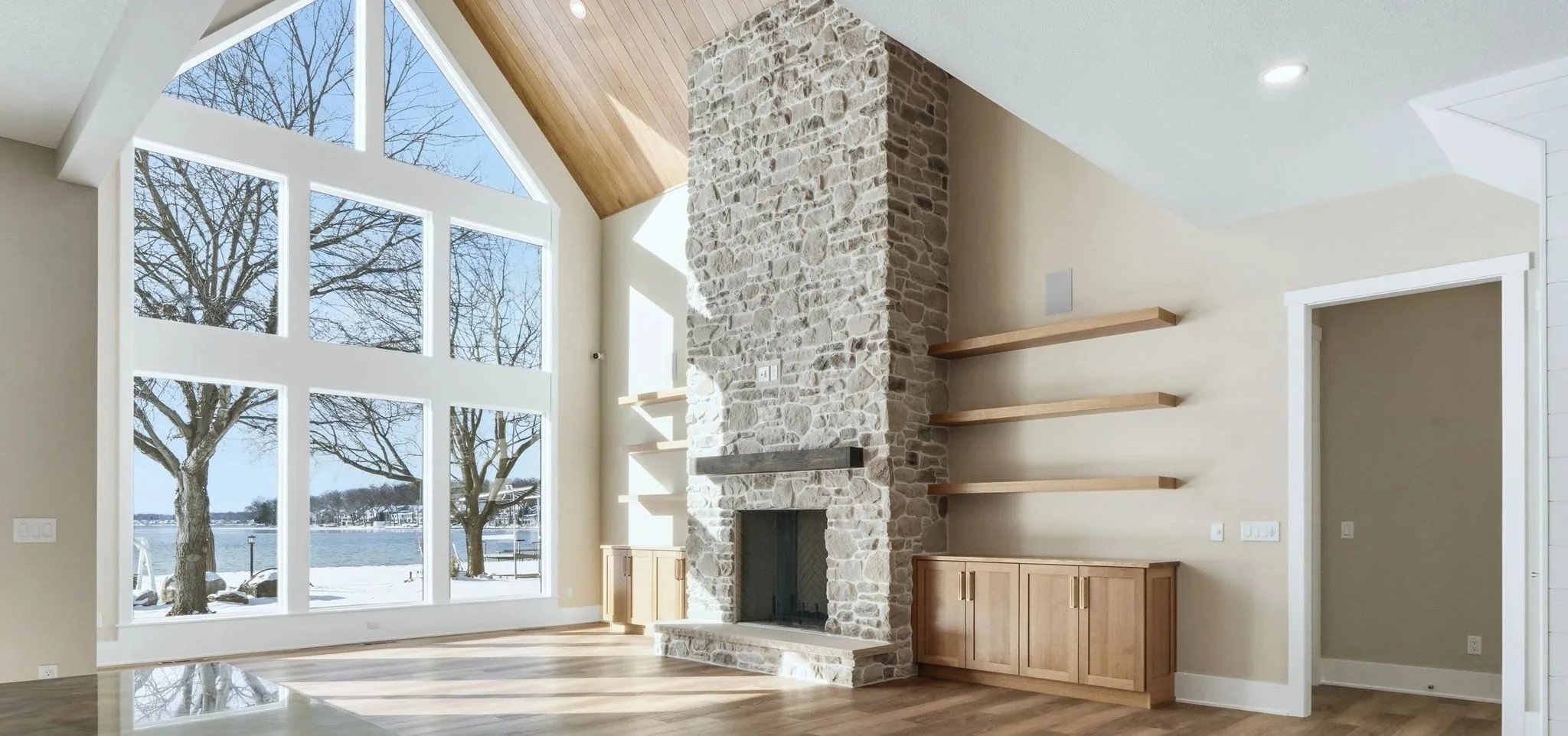 Living room with large window overlooking snowy landscape and water, stone fireplace with wooden shelves, and wooden cabinetry.