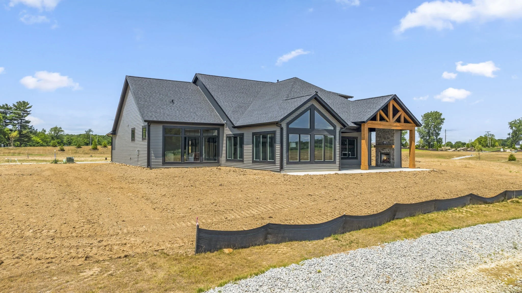 Newly built house with gray siding, large windows, and a covered porch with a stone fireplace, on a cleared lot with dirt and gravel.