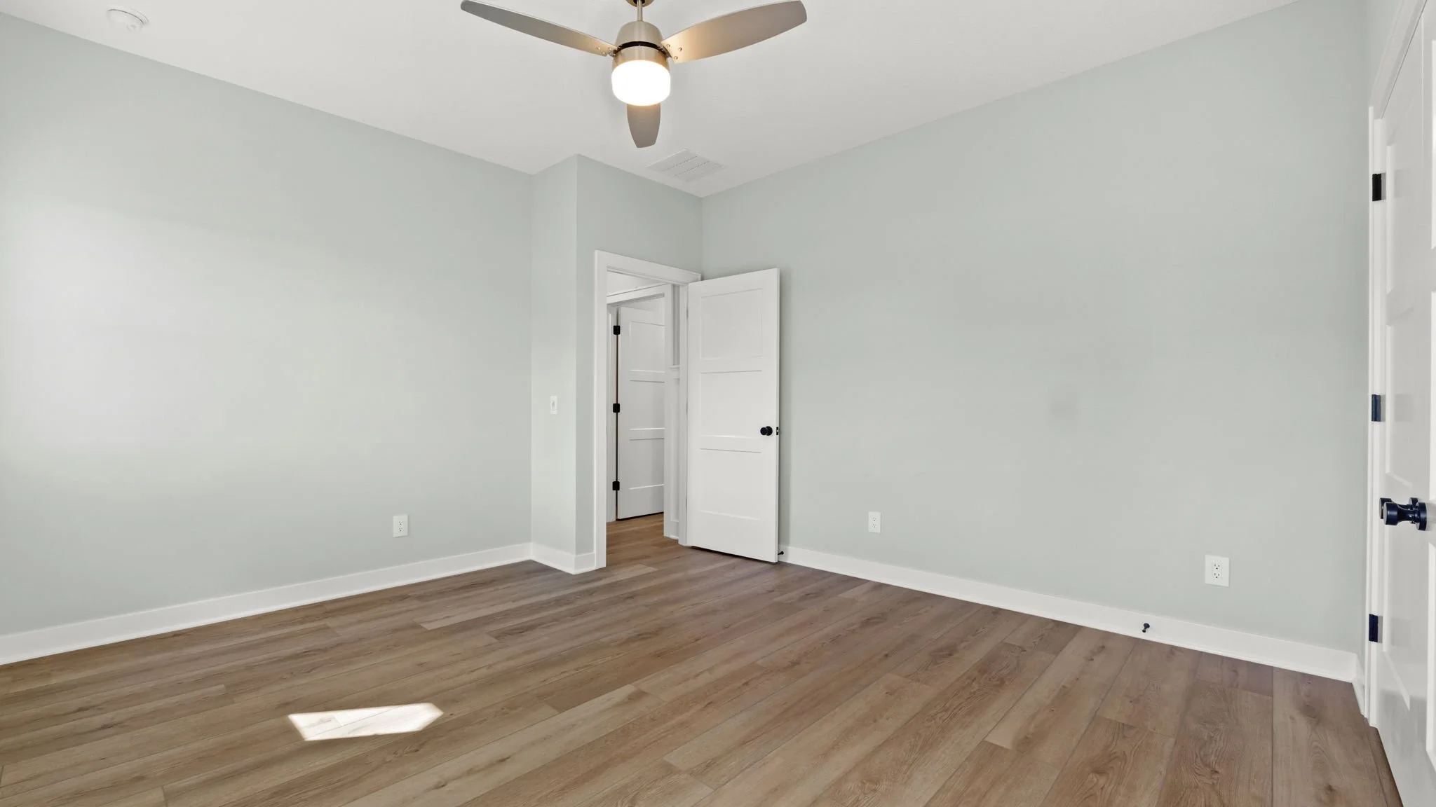 An empty room with light green walls, wooden flooring, two white doors, and a ceiling fan with a light.
