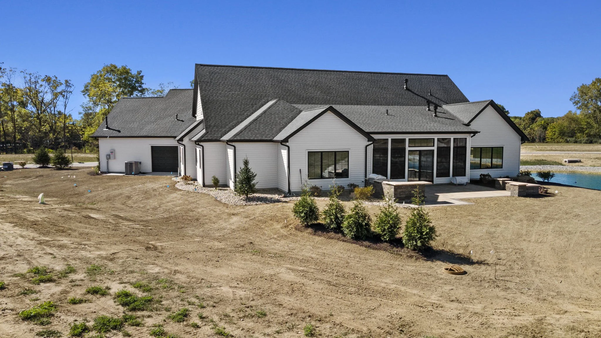 A modern house with white siding, a gray roof, large windows, glass doors, and a small patio in a yard with dirt and scattered bushes, surrounded by a few trees under a clear blue sky.