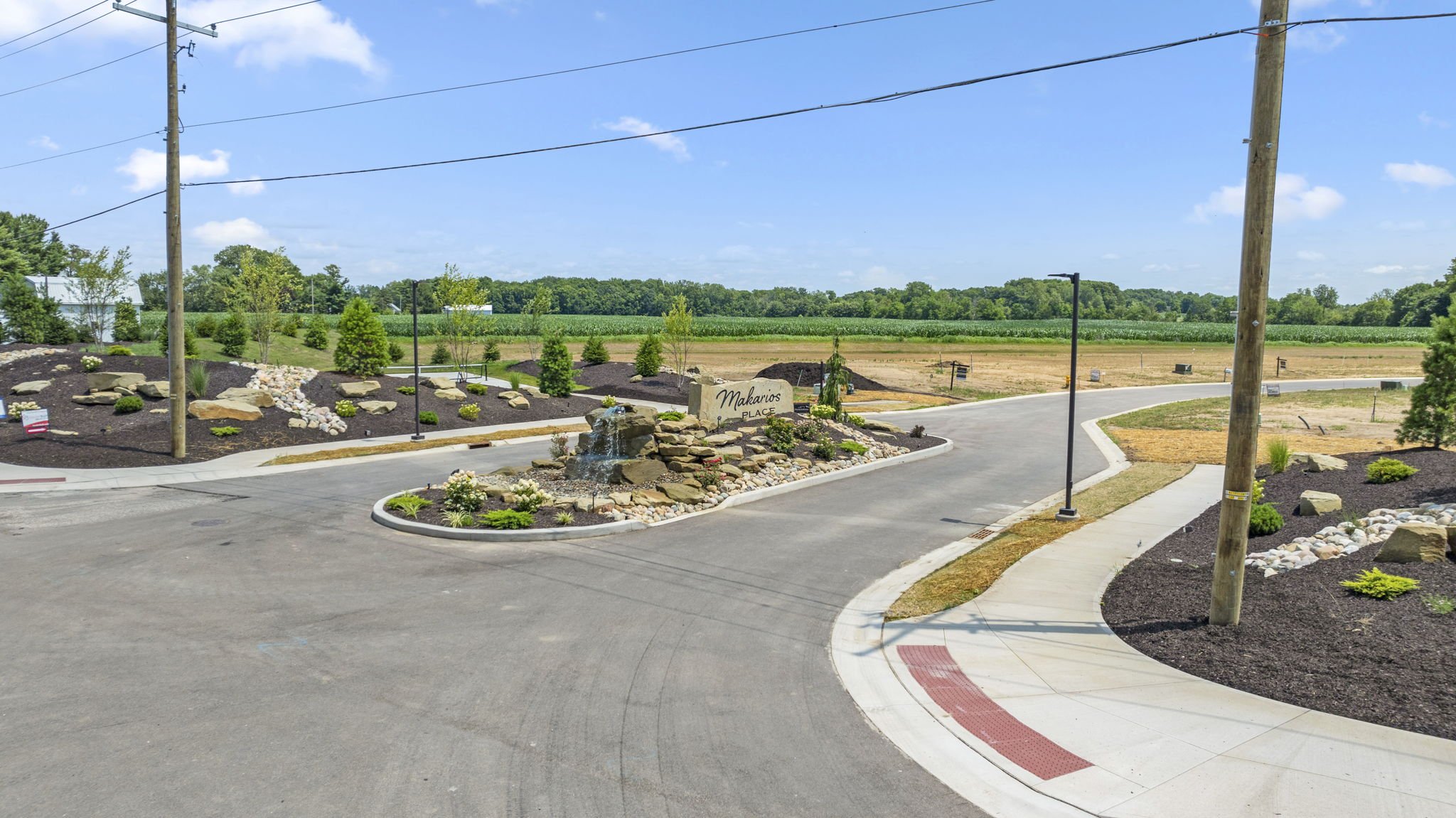 A landscaped roundabout at the entrance of a residential or commercial development, with a water fountain, small trees, rock features, and a sign reading "Makarios Place." In the background, there are fields and a clear blue sky with some clouds, along with utility poles and wires.