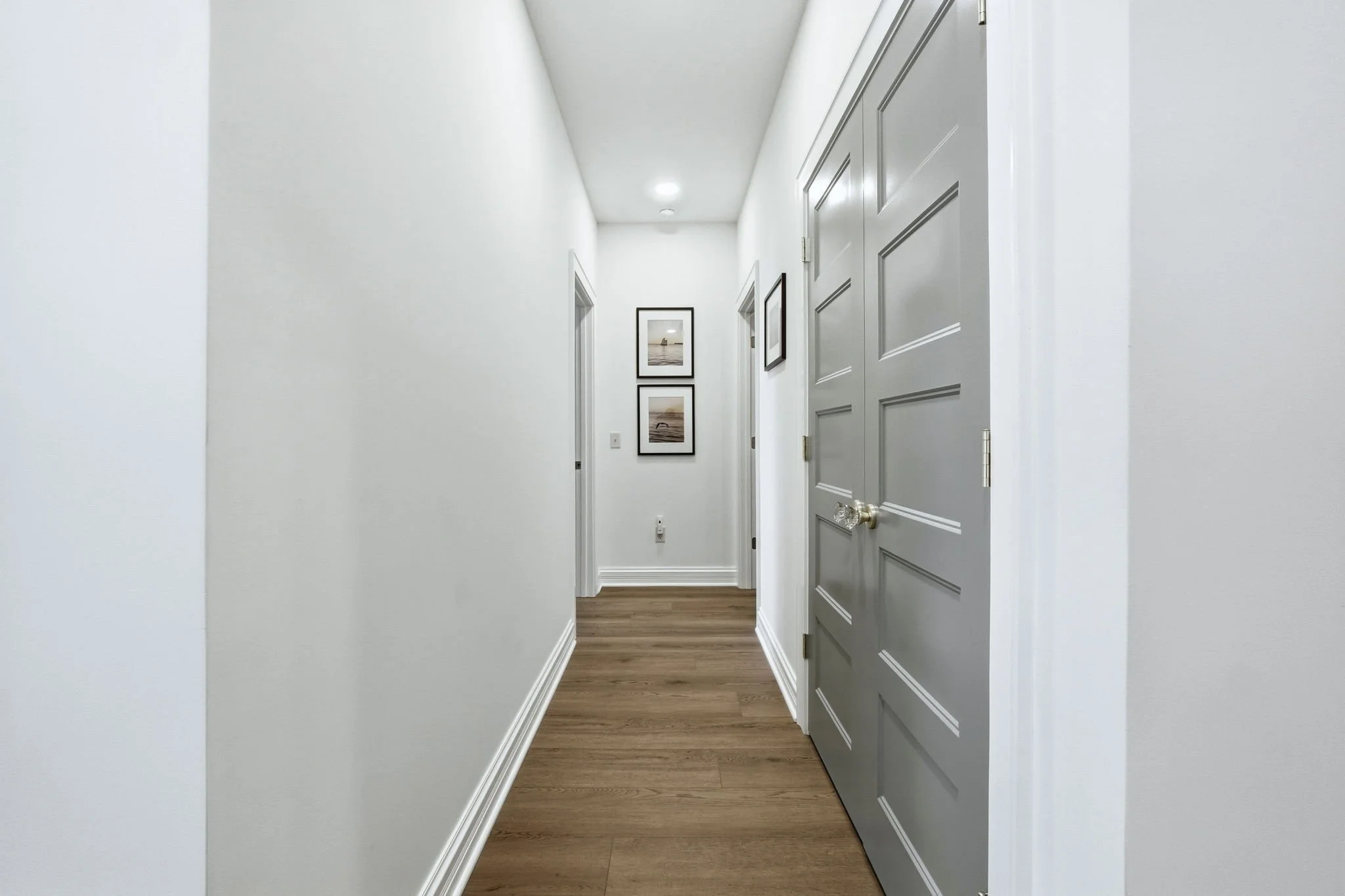 Empty hallway with white walls, wooden floor, gray doors on the right, and framed pictures on the wall at the end.