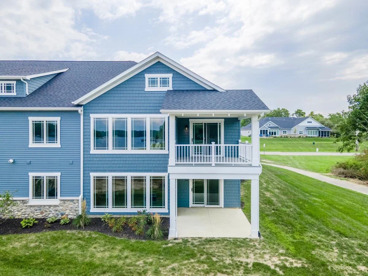 Blue multi-story house with large windows, a small balcony, and a backyard patio, surrounded by green lawn and neighboring houses under a partly cloudy sky.