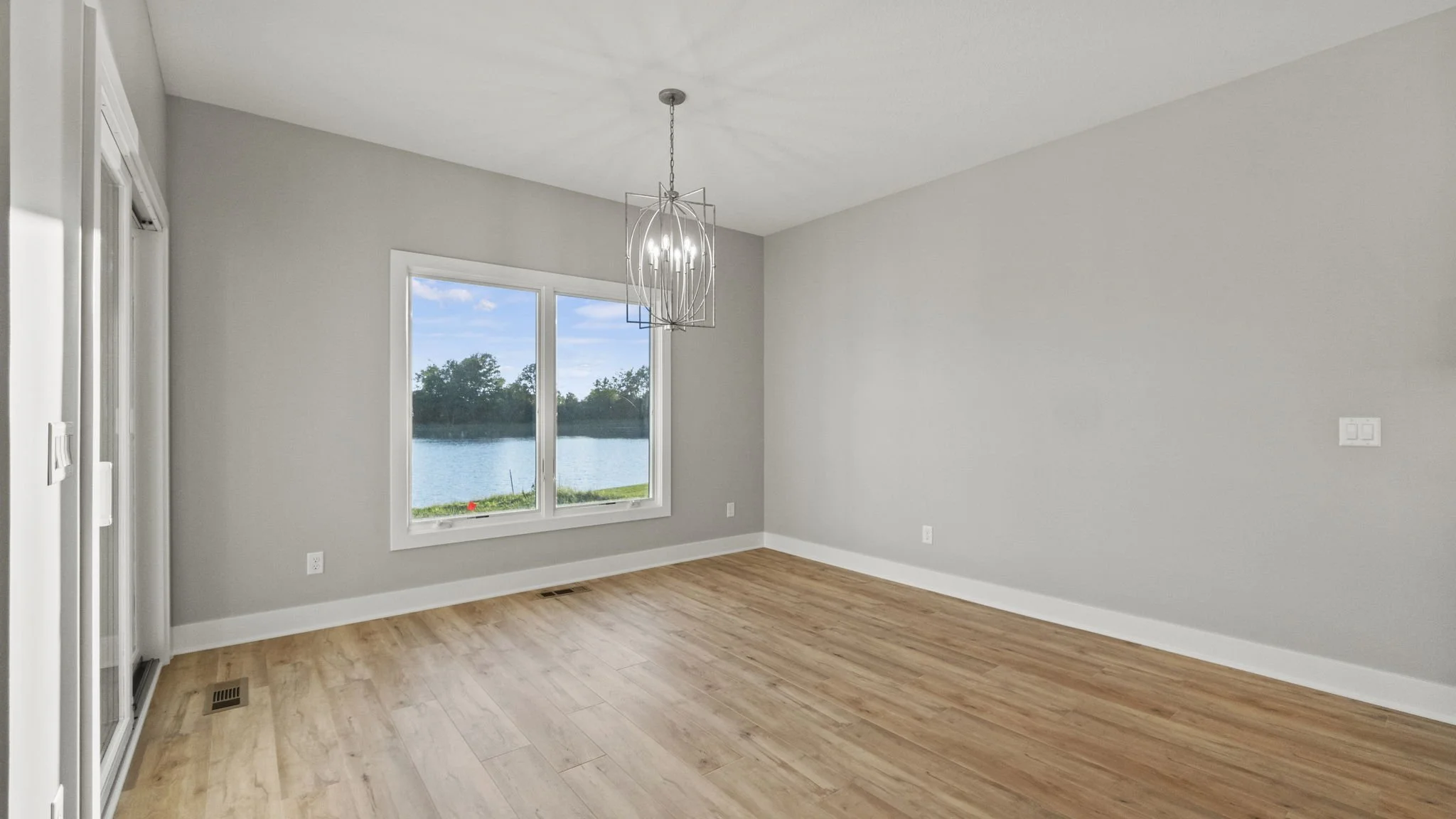 Empty room with hardwood floor, gray walls, a window showing a river view, and modern chandelier hanging from ceiling.