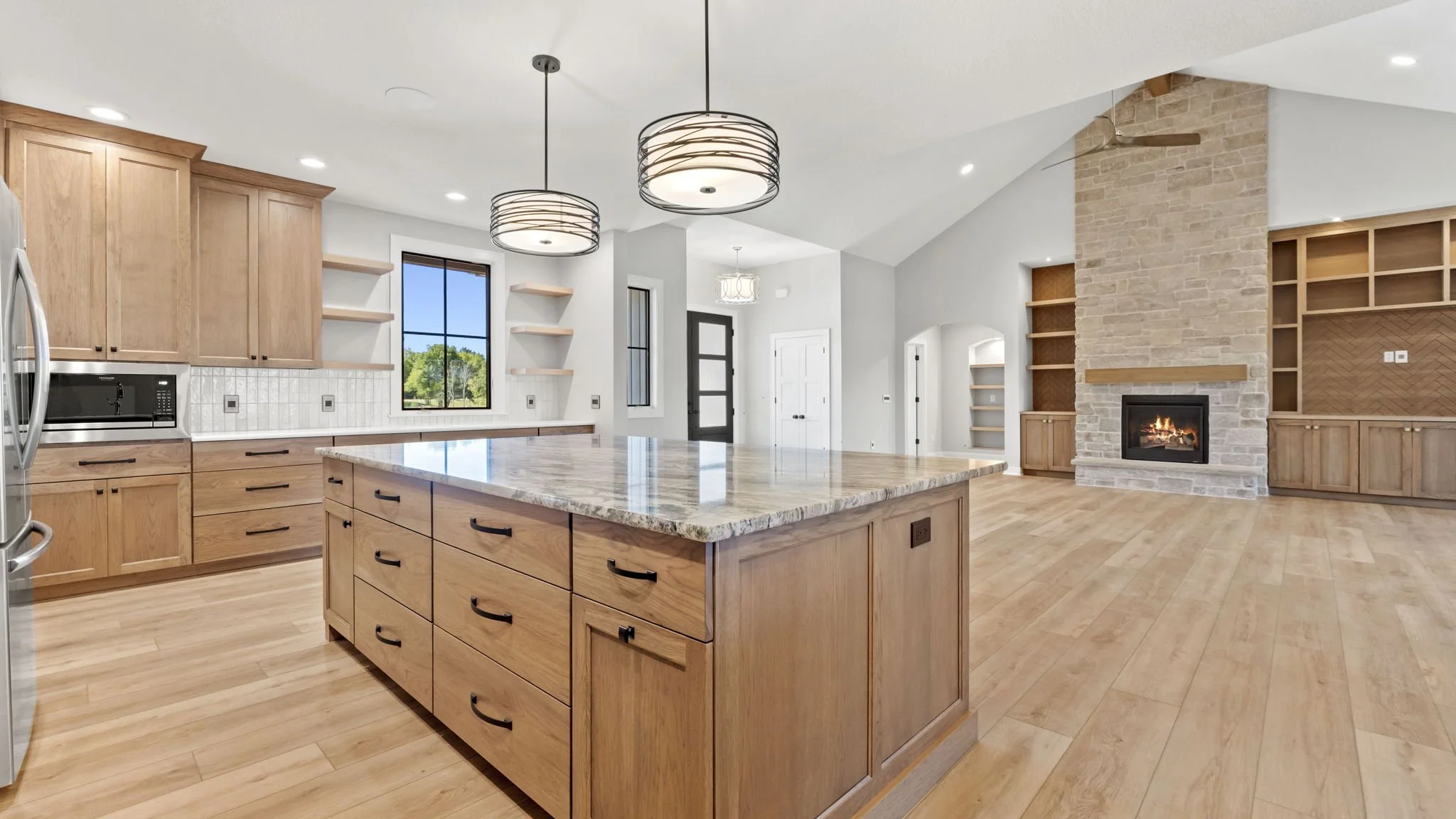 Open-concept kitchen and living room with wooden cabinetry, a kitchen island with marble countertop, hardwood floors, a stone fireplace, and built-in shelving. Large windows let in natural light.