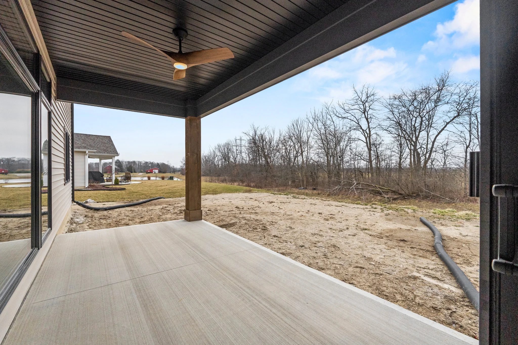 View from a covered porch with a ceiling fan, overlooking a backyard with grass, a pool, trees, and a cloudy sky.