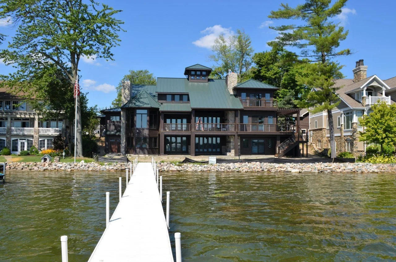 A large modern house with a dark exterior, multiple windows, and a green metal roof, situated on a lakeside with a dock extending into the water, surrounded by trees and neighboring houses under a blue sky.