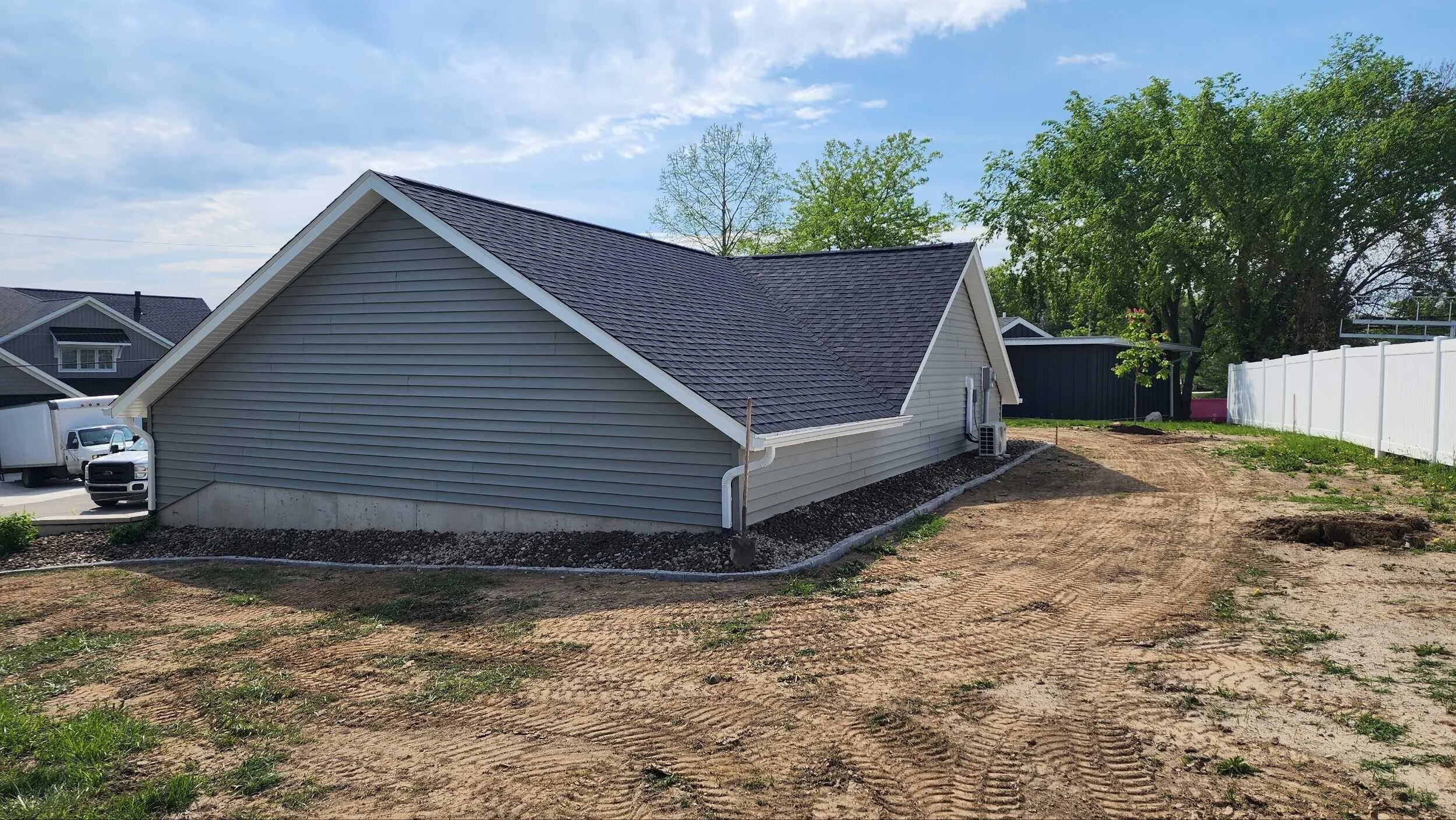 Side view of a newly built house with vinyl siding, a dark shingle roof, and a white gutter system. The yard is under construction, with dirt and sparse vegetation, and a white fence borders the property. In the background, there are green trees, oth