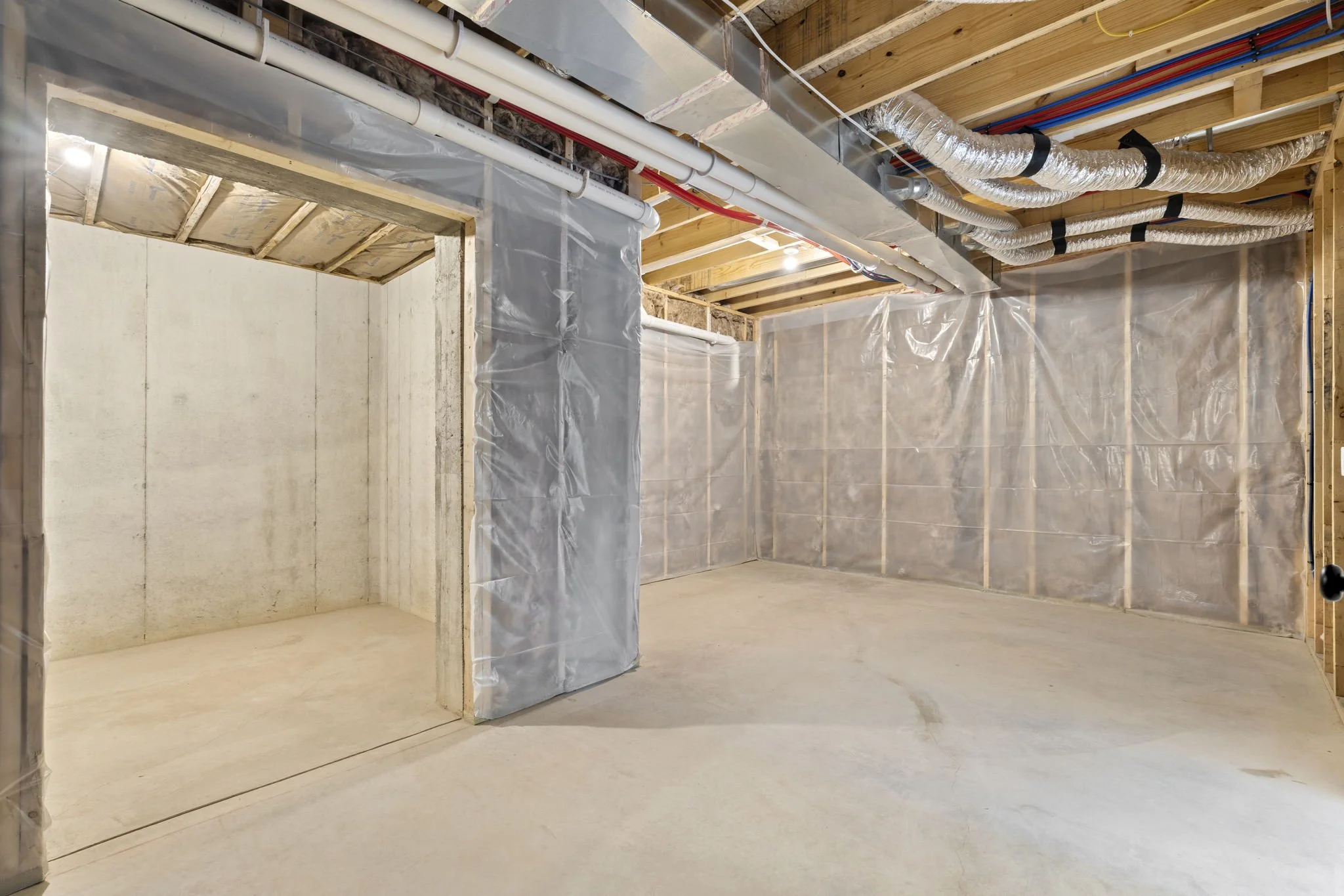 Empty basement with unfinished walls, exposed wooden ceiling beams, exposed ducts and pipes, and plastic sheeting on walls.
