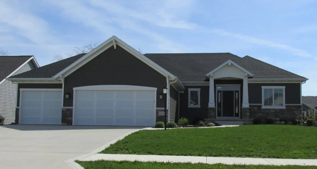 A modern, single-story house with a gray exterior, white trim, and a three-car garage, situated on a green lawn under a clear blue sky.