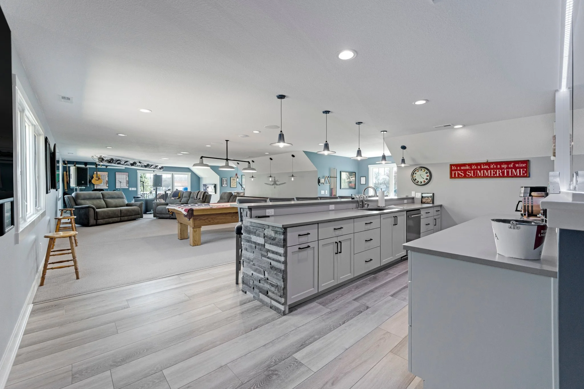 Open-concept living room and kitchen with gray cabinets, a bar counter, pendant lights, and a pool table, decorated with music and beverage-themed decor.