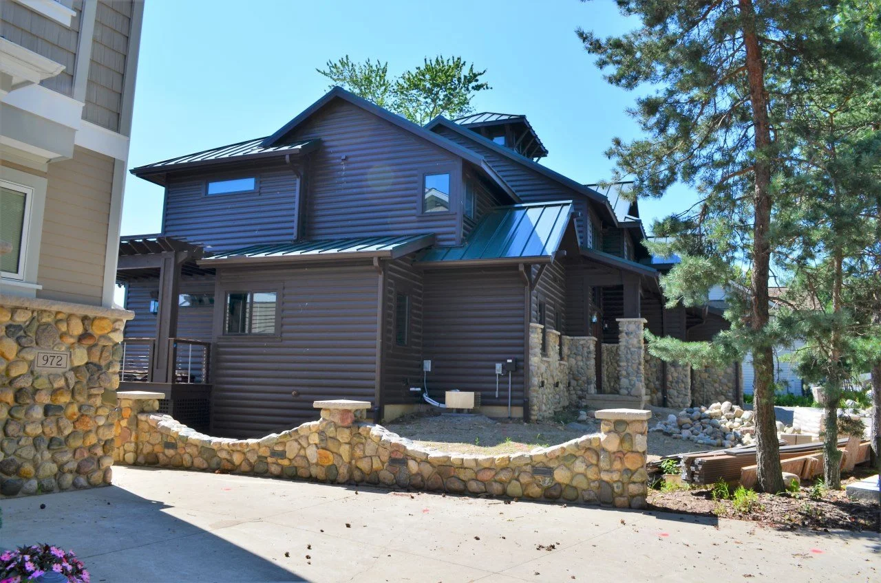 Side view of a modern dark brown house with a metal roof, stone columns, and a stone wall boundary. There are tall trees nearby and construction materials on the side.