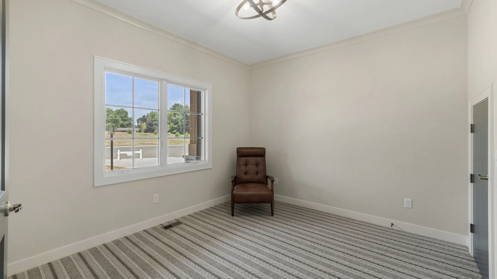 Empty room with beige walls, a brown armchair near a window, striped carpet, and a modern ceiling light fixture.