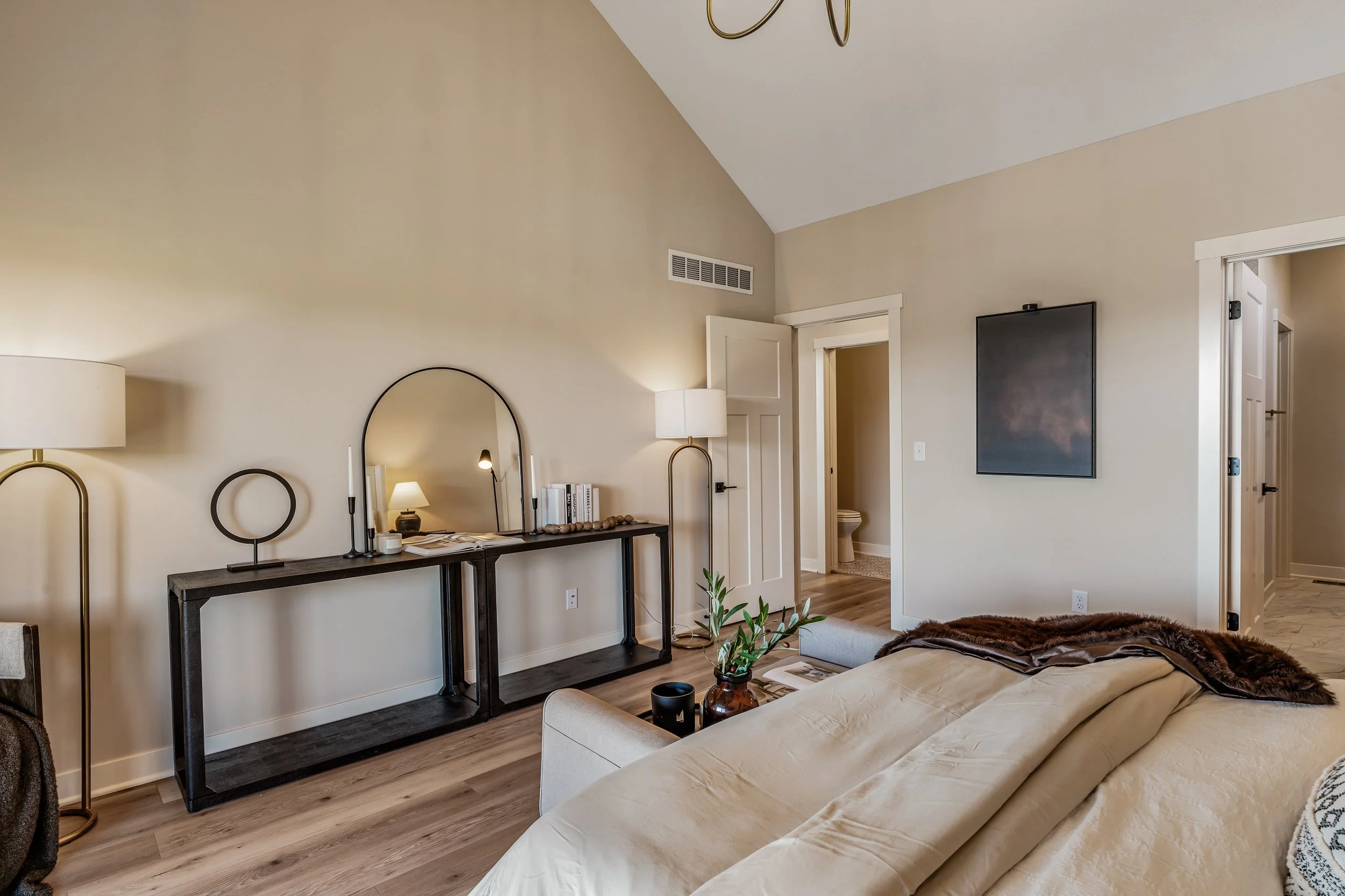 Modern bedroom with beige walls, a bed with beige duvet, a black console table with a mirror, books, and decor, and a wall-mounted TV.