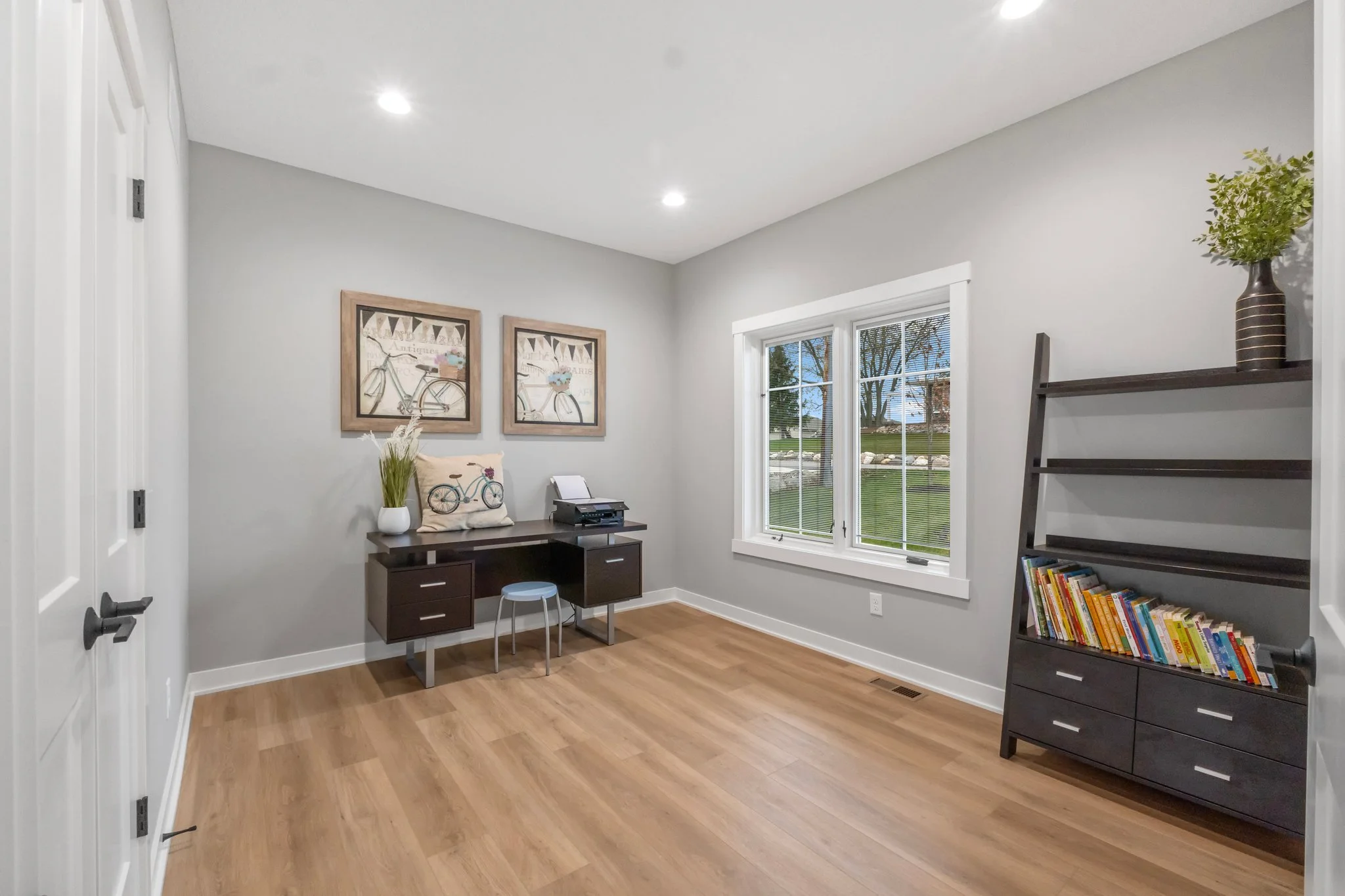 Empty room with hardwood flooring, light gray walls, a window showing a green outdoor area, a dark brown bookshelf with books, a small dark desk with a printer, decorative art of bicycles on the wall, and a vase with greenery on the bookshelf.