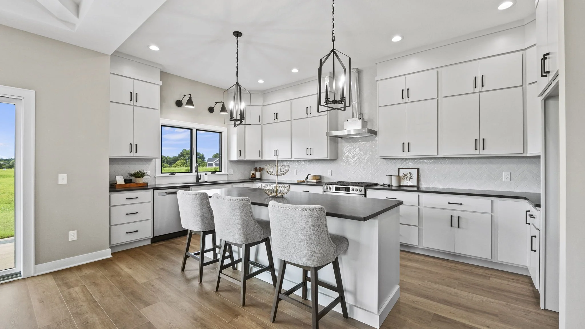 Modern kitchen with white cabinets, black handles, a gray island with barstools, black pendant lights, and a window overlooking a green field.