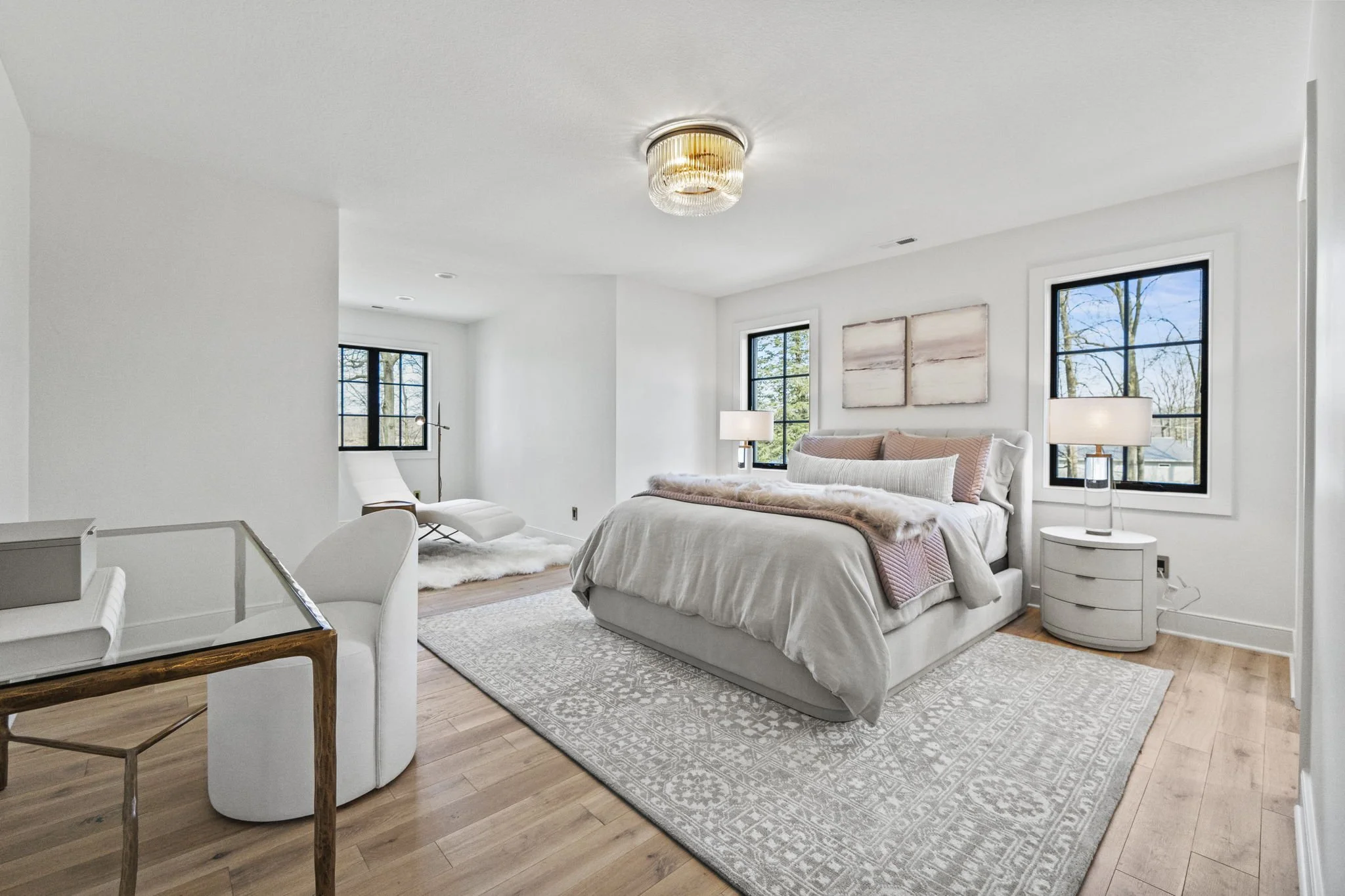 Bright, modern bedroom with white walls, light wood flooring, and large black-framed windows showing trees outside. The room features a large bed with white and pink bedding, two matching bedside tables with lamps, a white armchair, an art piece divi
