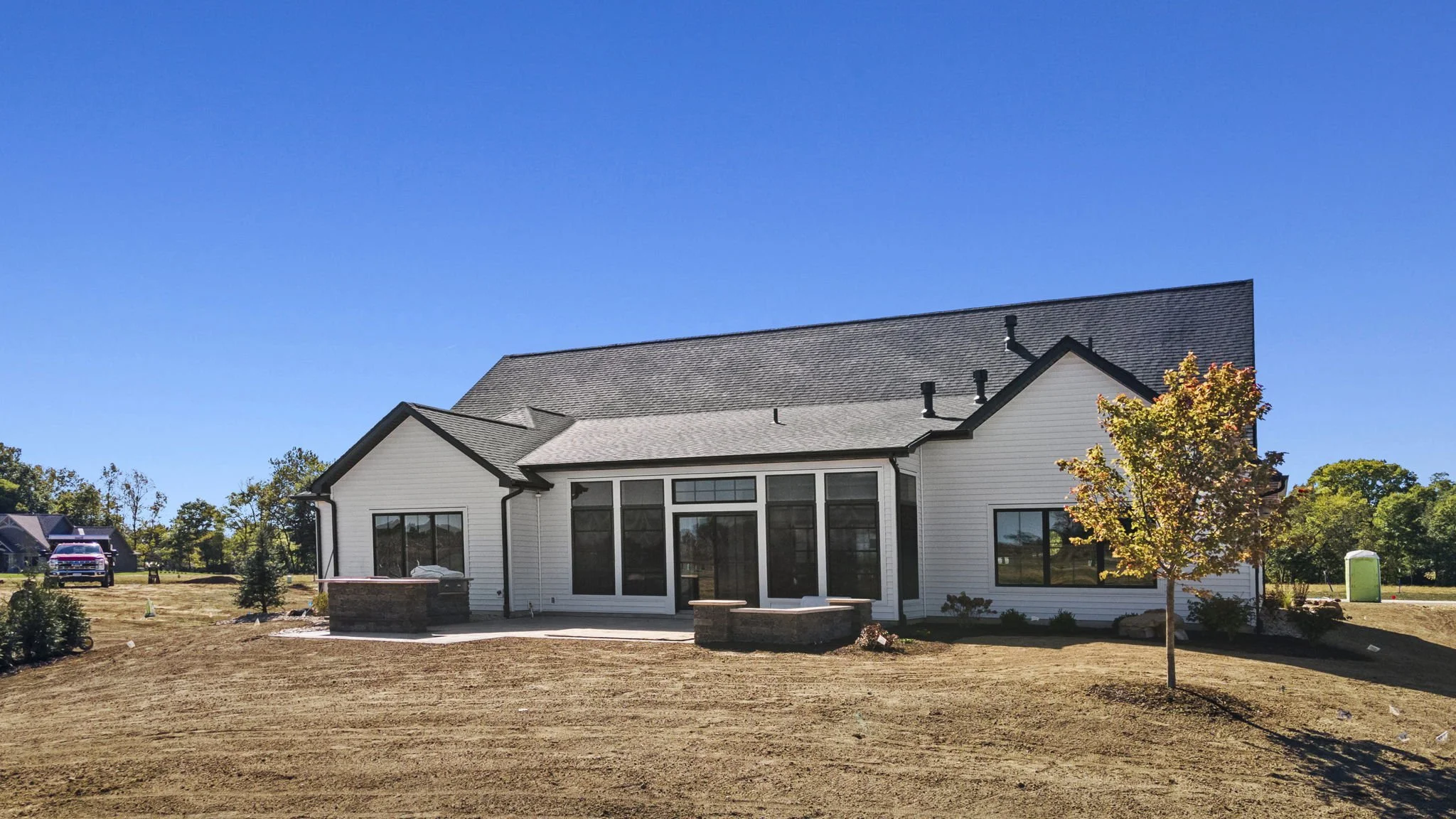 A newly constructed house with white siding, black window frames, and a gray shingled roof. It has large windows on the back side and a small tree with fall foliage in the yard. The ground is bare, likely due to recent landscaping, with a clear blue 