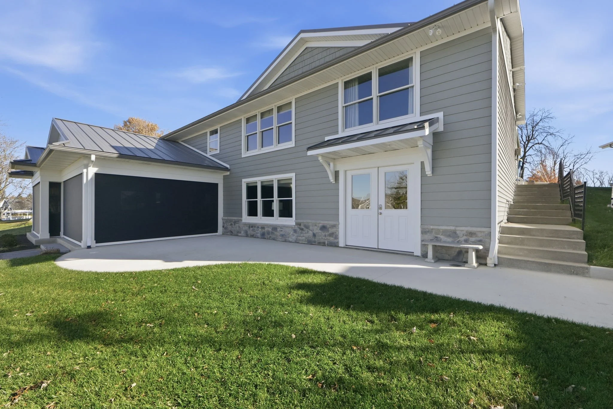 A modern two-story house with gray siding, white trim, and a stone foundation, featuring a concrete patio, stairs leading to the yard, and a blue sky background.